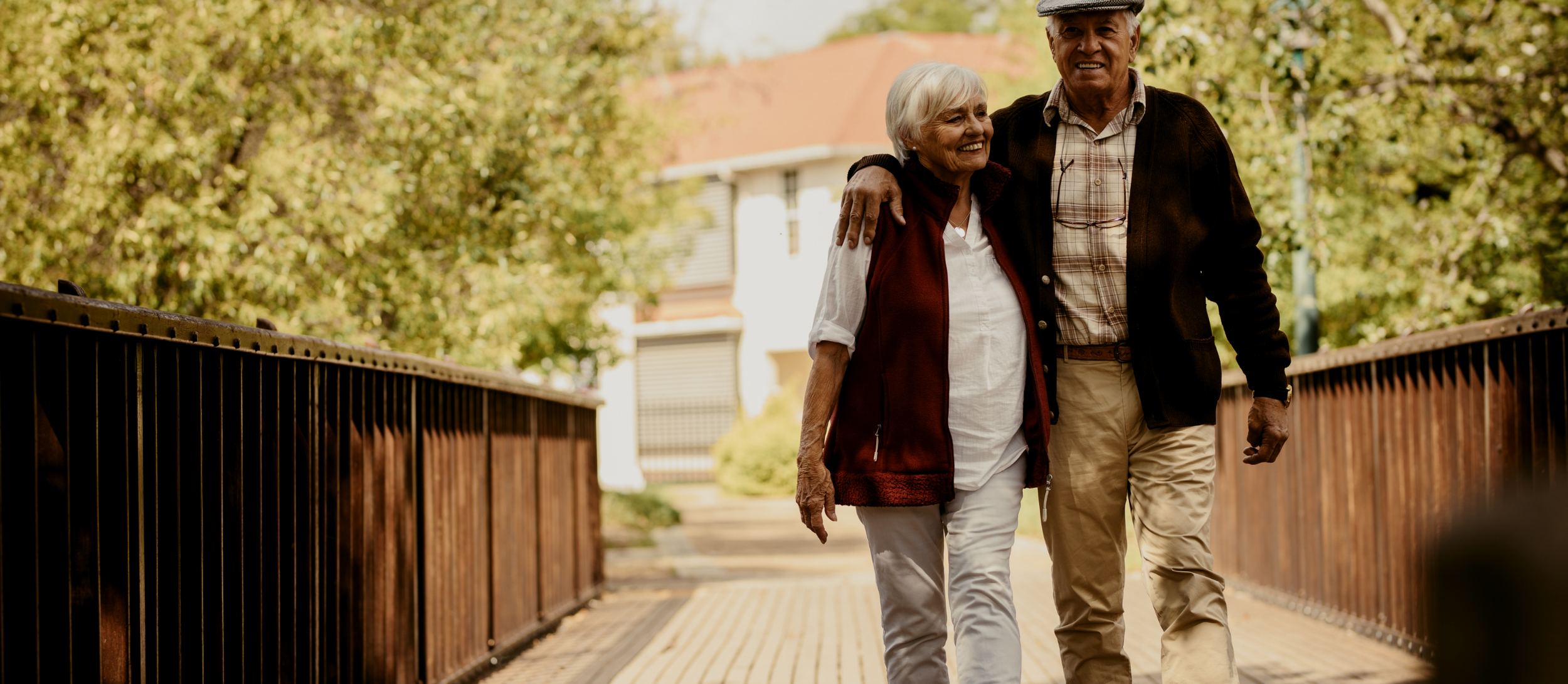 An elderly couple walking arm-in-arm on a wooden bridge outdoors, smiling and enjoying each other's company, surrounded by trees and a house in the background.
