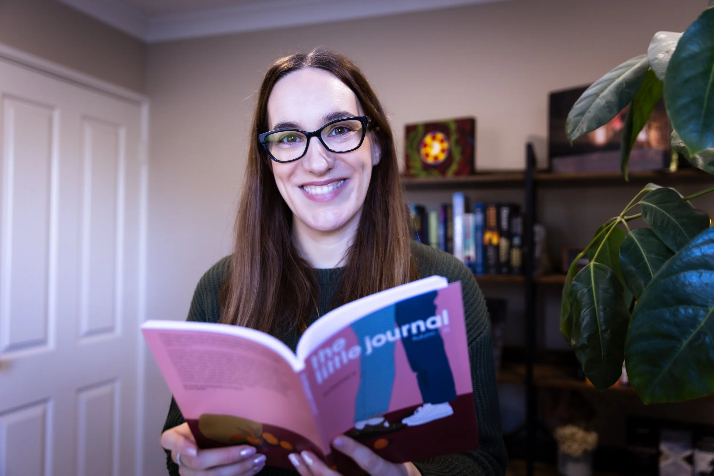 A photo of Britt McCarthy smiling and holding up a copy of the Little Journal. She has brown hair and black glasses