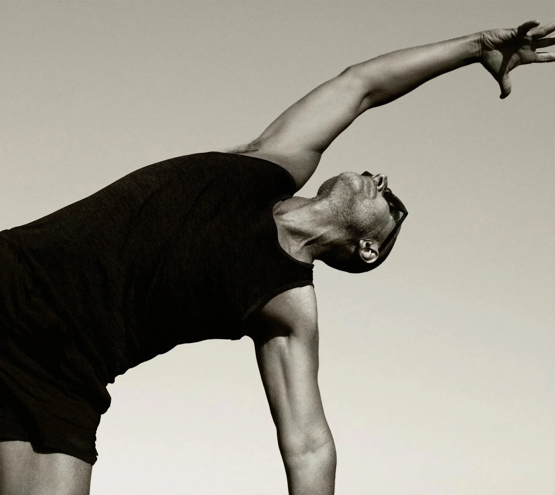 A man wearing glasses and black clothing, in a yoga pose on a plain background, with his head tilted back and arm extended upward.