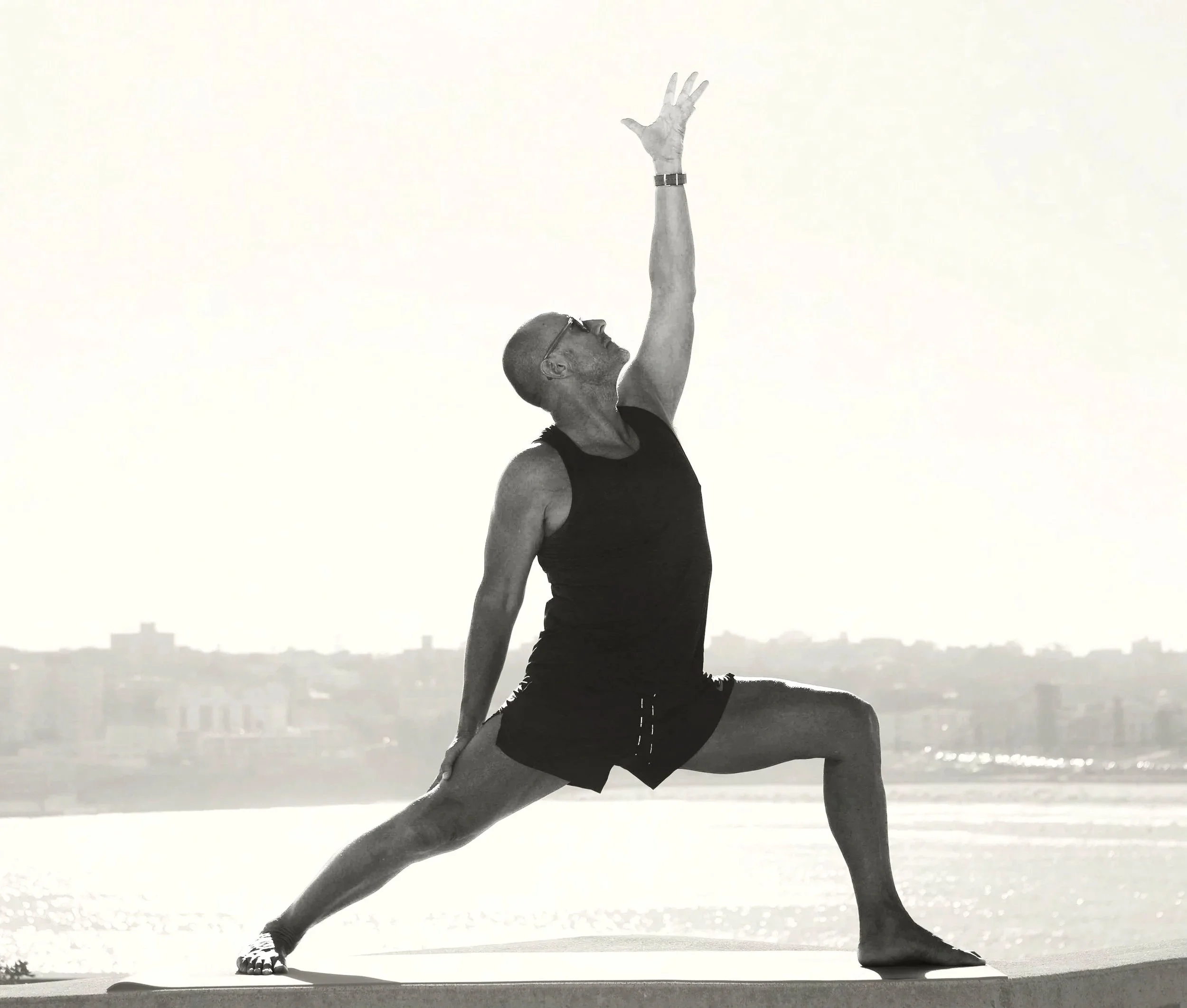 A man practicing yoga outdoors, performing a side stretch pose with one arm reaching upward and the other hand touching his ankle, near the water with buildings in the distant background.