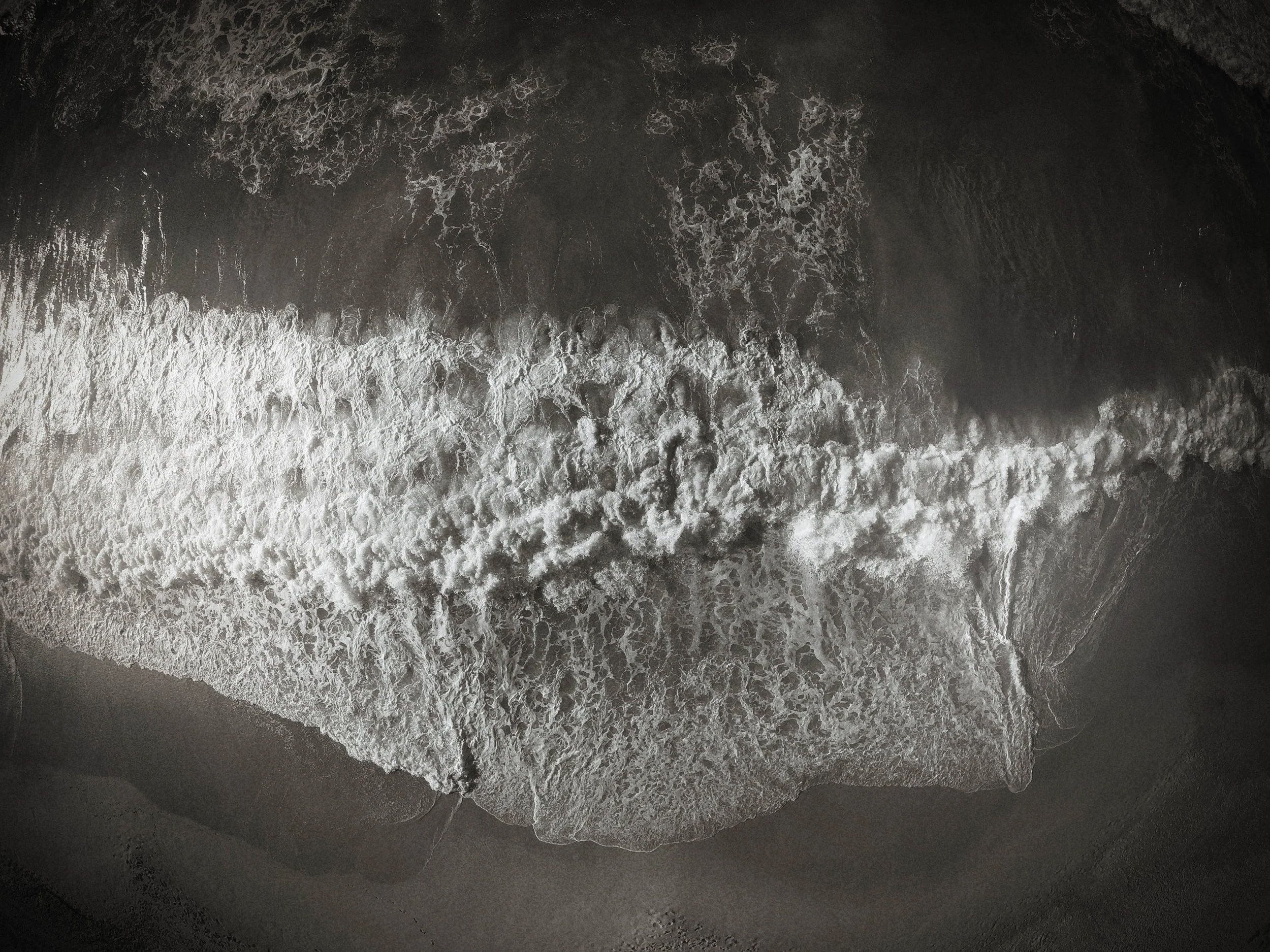 Waves crashing onto a dark sandy beach from an aerial view at sunset or sunrise.