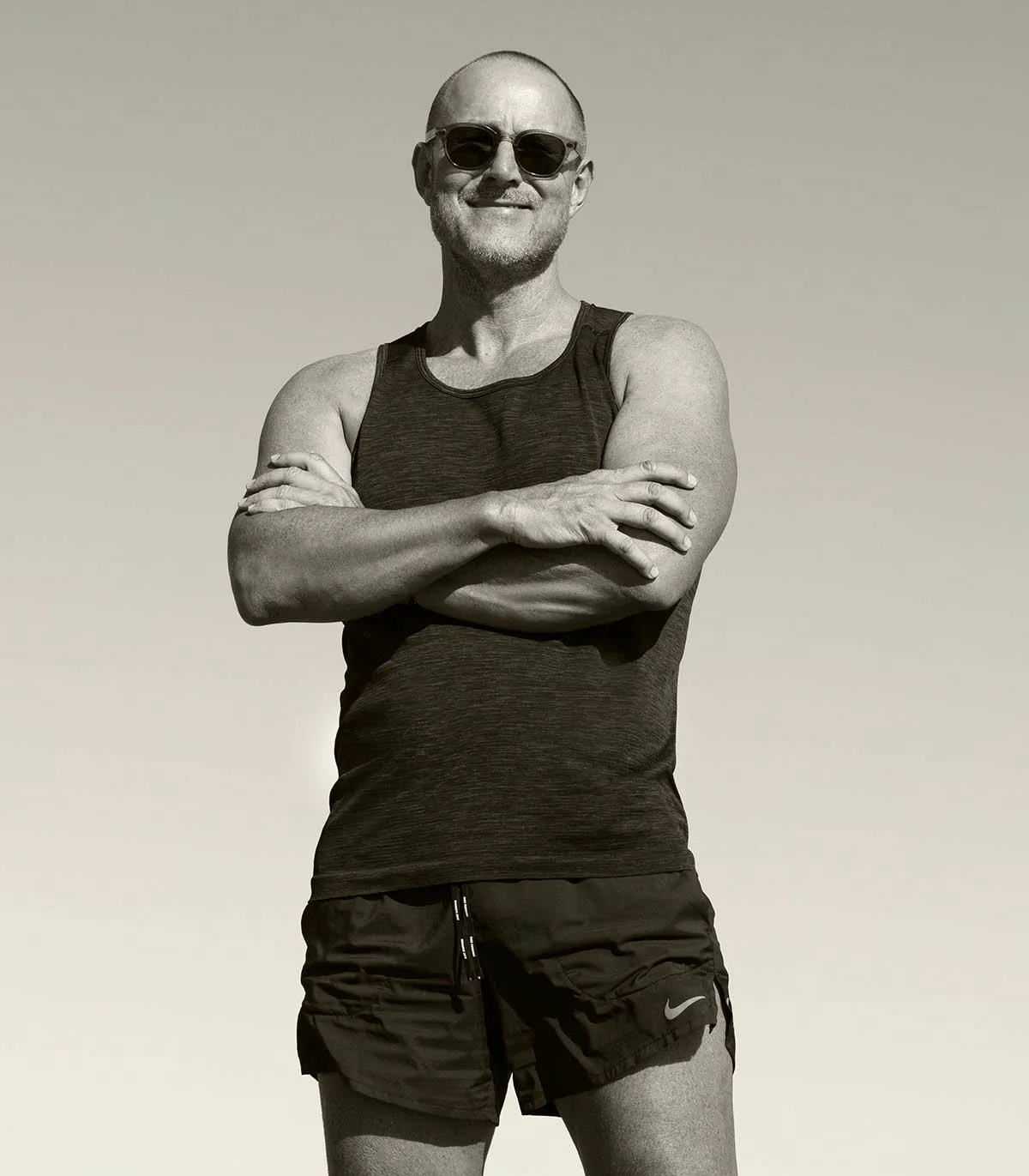 A smiling man wearing sunglasses, a black athletic tank top, and black Nike shorts, standing with arms crossed against a clear sky.