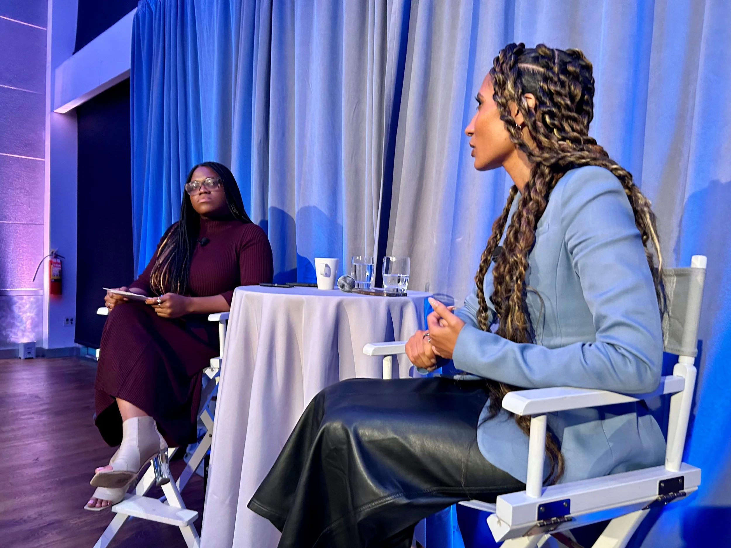 Two women participate in a panel discussion onstage, seated beside a table with microphones, water glasses, and cups.