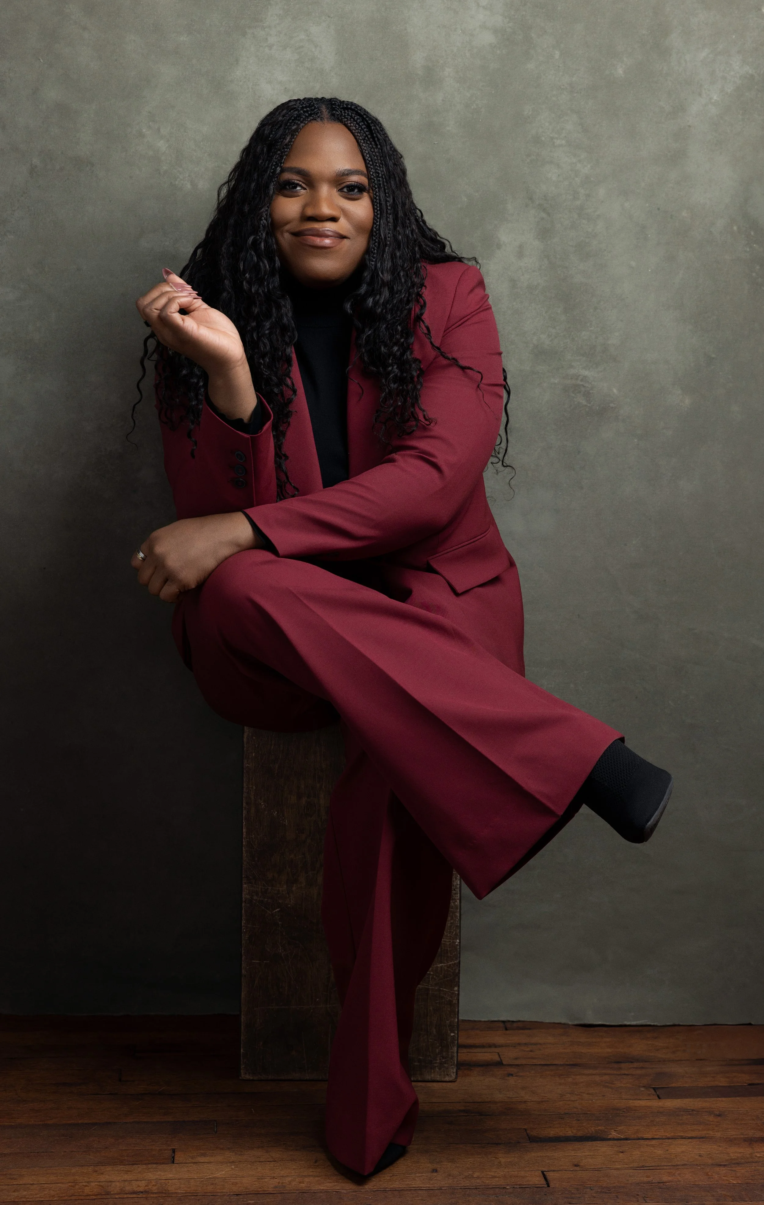 Professional Black woman in a burgundy suit and black top, seated against a textured gray wall.