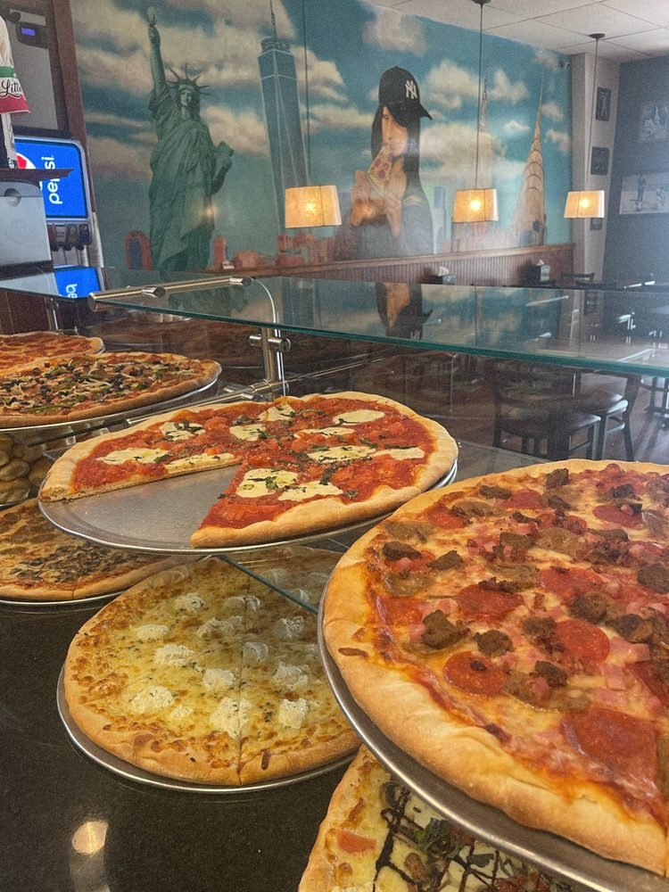 Several different pizzas on display on glass shelves in a restaurant, with a mural of the Statue of Liberty, a woman wearing a New York Yankees cap, and city buildings in the background.