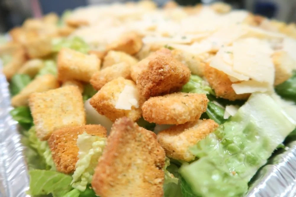 Close-up of a Caesar salad with croutons and shaved cheese in a foil container.