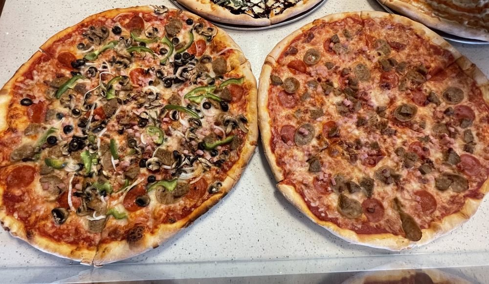 Two large pizzas on a white surface. The pizza on the left has toppings including black olives, green peppers, sausage, onions, and possibly ham or bacon. The pizza on the right has pepperoni and sausage as toppings.