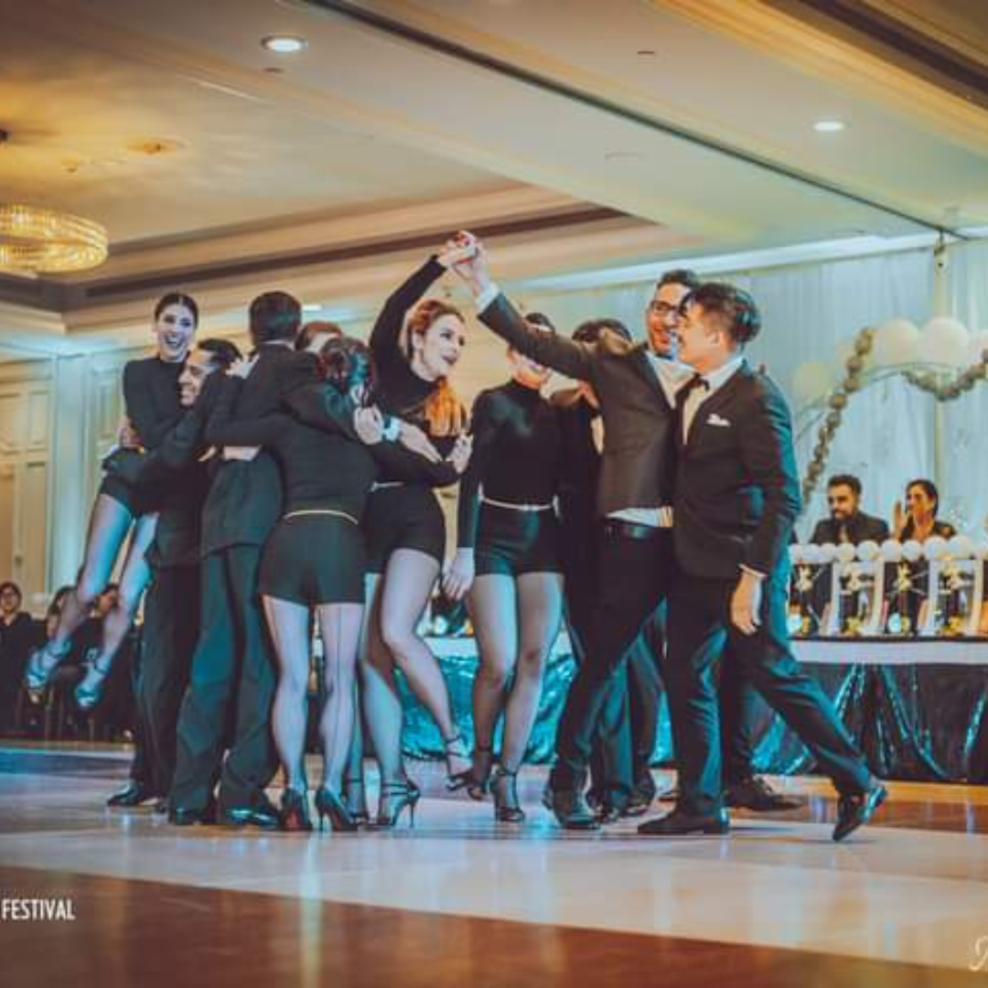 Group of people dancing and celebrating at an indoor event or party, with women dressed in black outfits and men in suits, on a wooden dance floor.