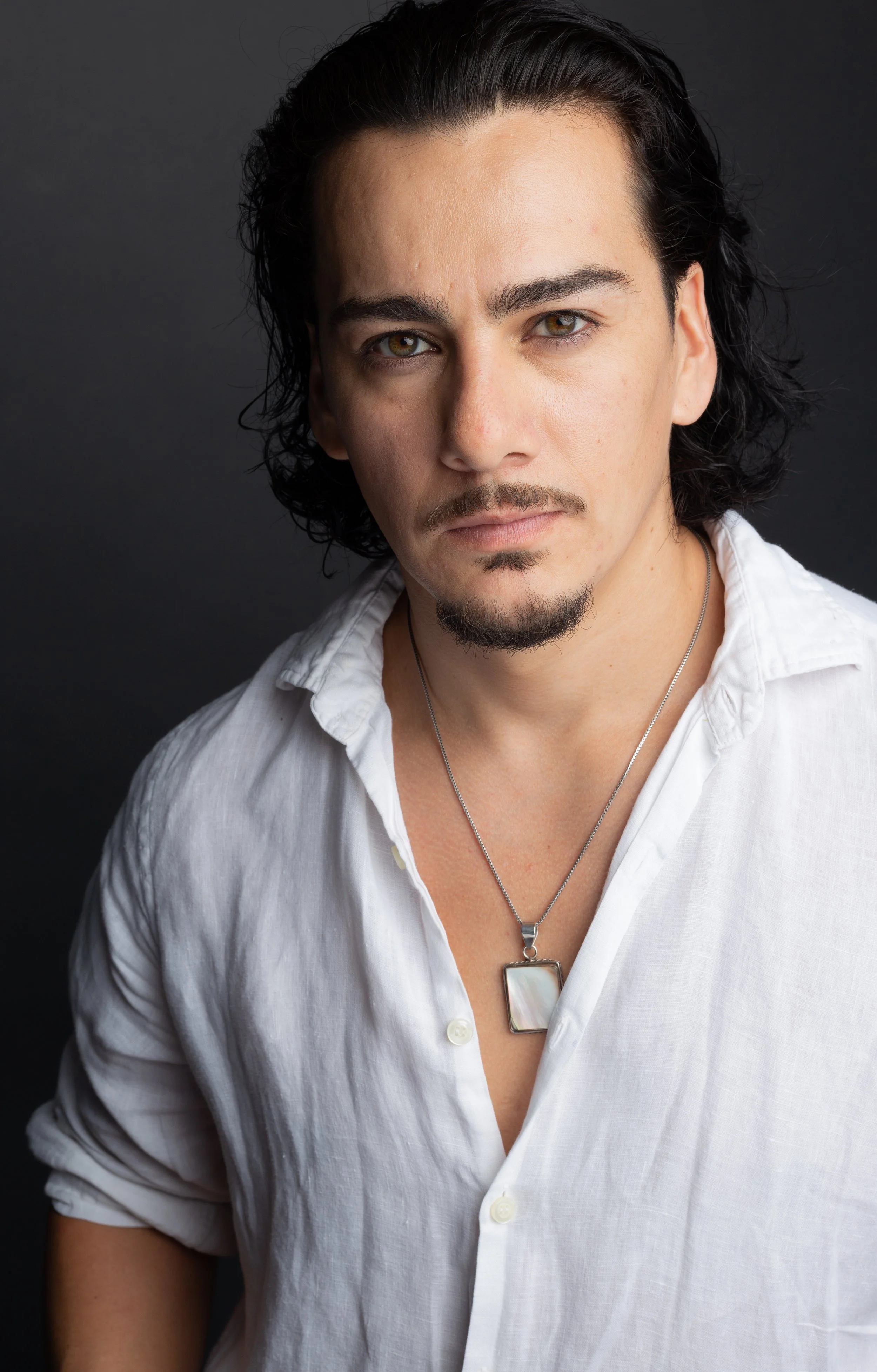 A man with shoulder-length black hair, brown eyes, and a goatee, wearing a white button-up shirt slightly unbuttoned, with a necklace featuring a rectangular pendant, against a dark background.