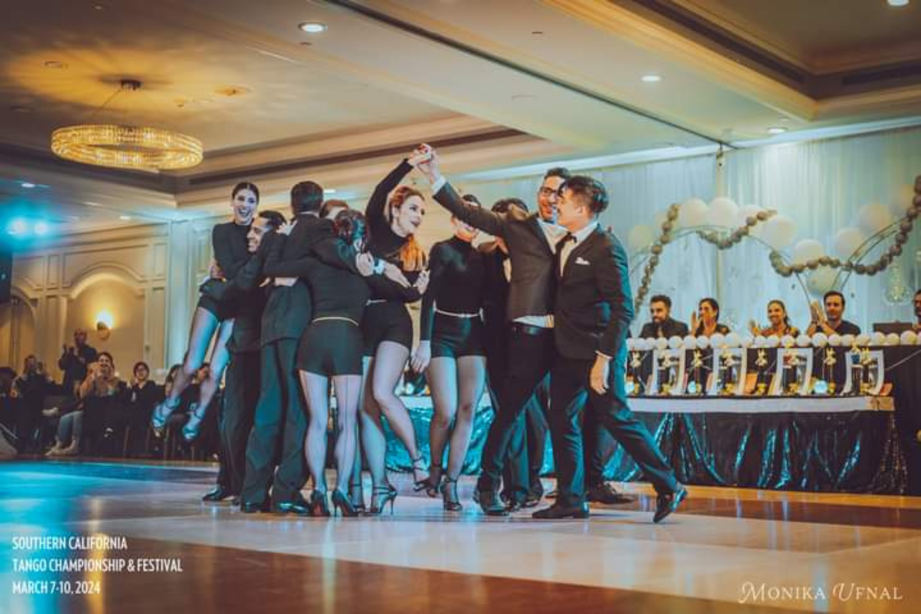A group of people in formal attire participating in a dance at the Southern California Tango Championship & Festival, March 7-10, 2024, with some onlookers and judges seated at a table decorated with balloons and banners.