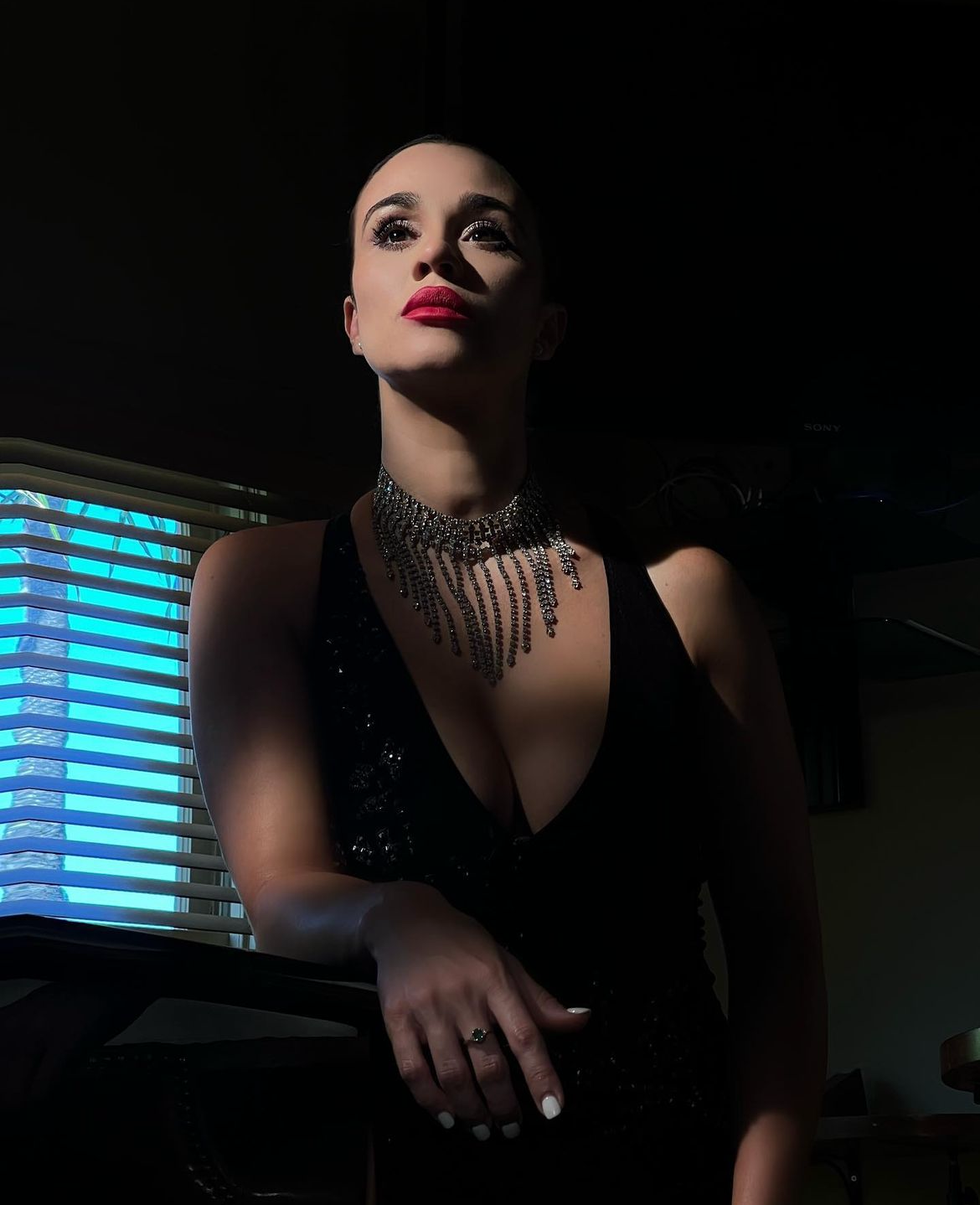 A woman in glamorous attire with a black dress and sparkly necklace, standing near a neon-lit window, with dramatic lighting highlighting her face and upper body.