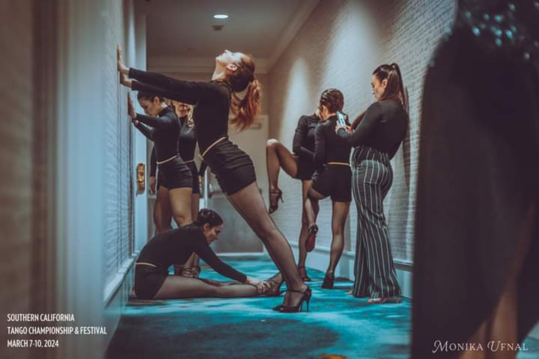 Women dressed in black dance costumes and heels demonstrate tango moves in an indoor hallway. The woman in the foreground leans back with arms extended, while another woman to the right is seated, adjusting her shoe. A woman in striped pants stands nearby, watching. The setting is for the Southern California Tango Championship & Festival, held from March 7-10, 2024, with the photographer credited as Monika Ufnal.