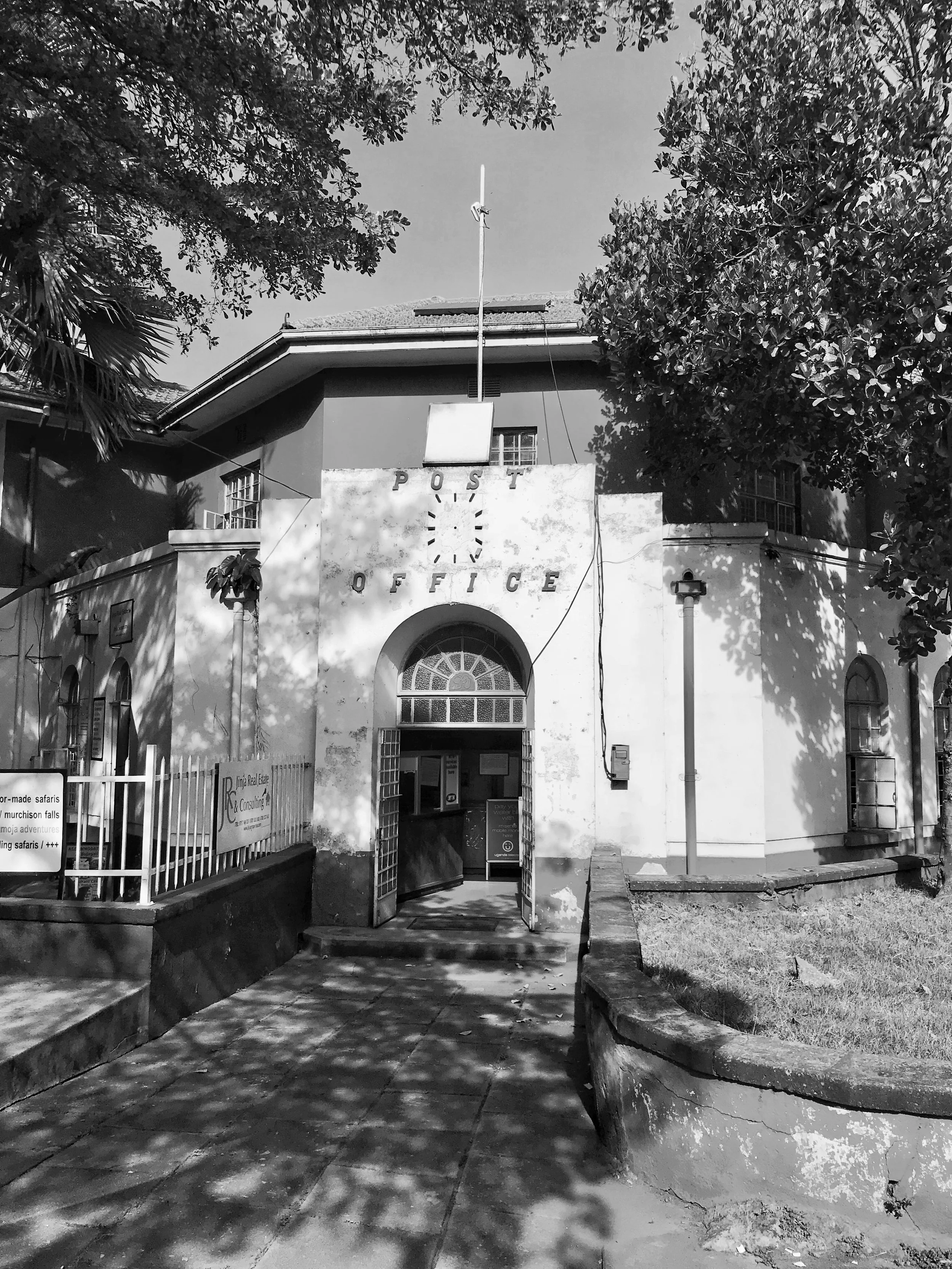 A black-and-white photograph by Jim Joel of the historic post office in Jinja, eastern Uganda. The building features a distinctive arched entrance and a sign bearing the word “POST,” framed by trees on either side. 