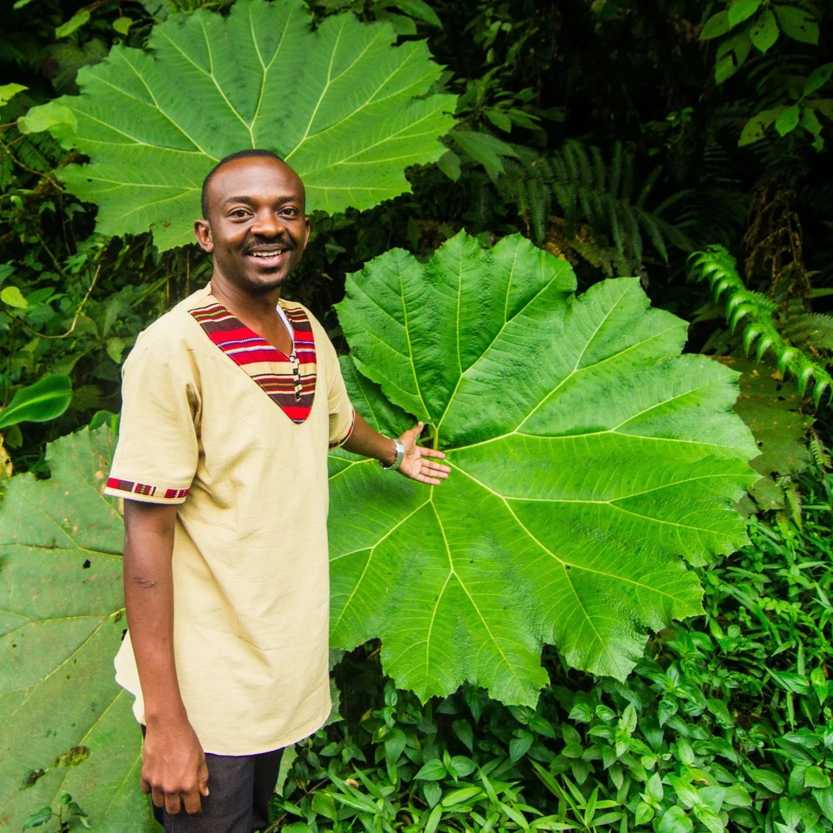A man standing outdoors in front of large green leaves and dense foliage, smiling and gesturing with his right hand.
