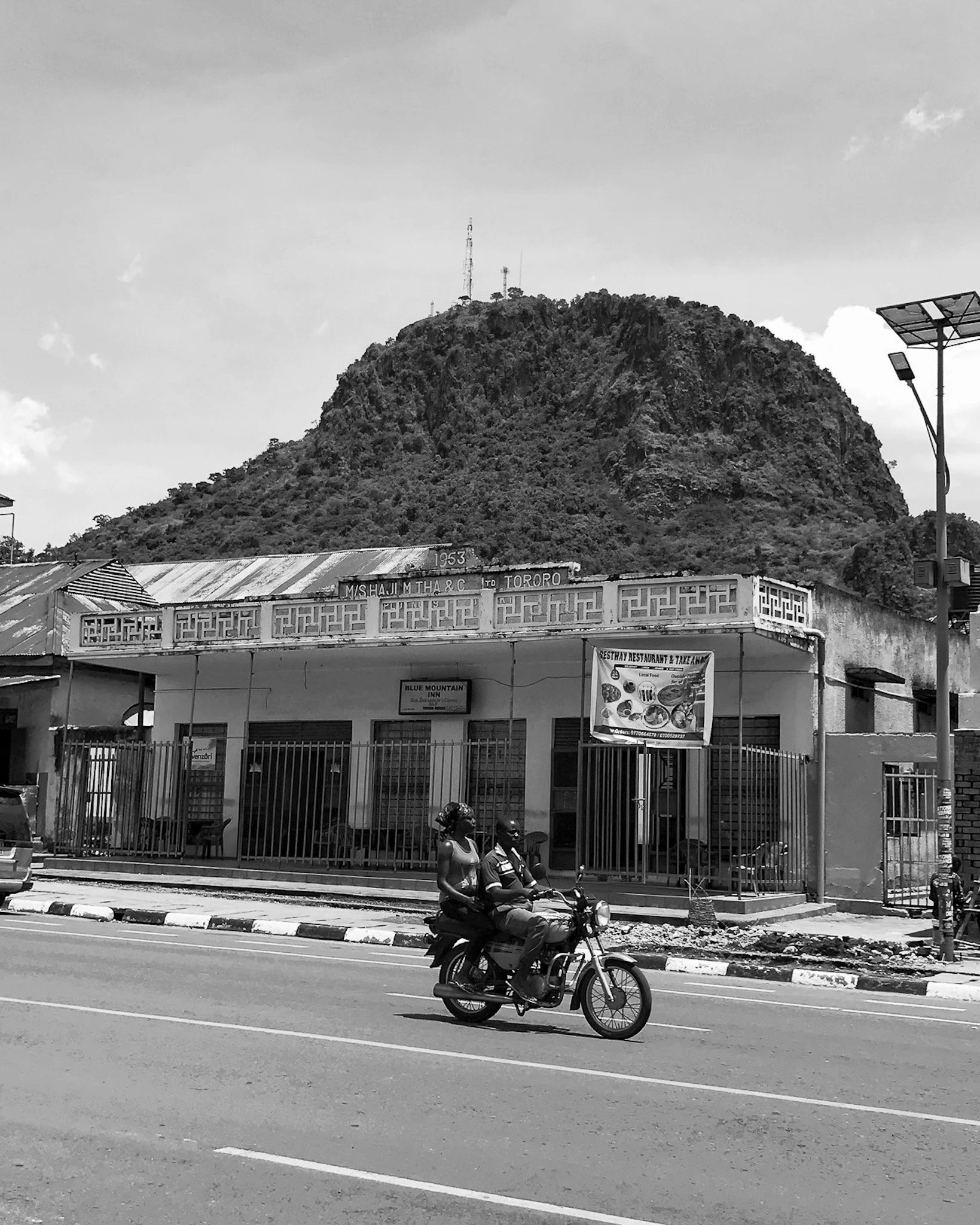A street scene showing two people riding a motorcycle along the road. In the background, there's a building with a sign that reads 'Blue Mountain Inn' and a mountain with communication towers on top. There's also a person walking and some street lamp