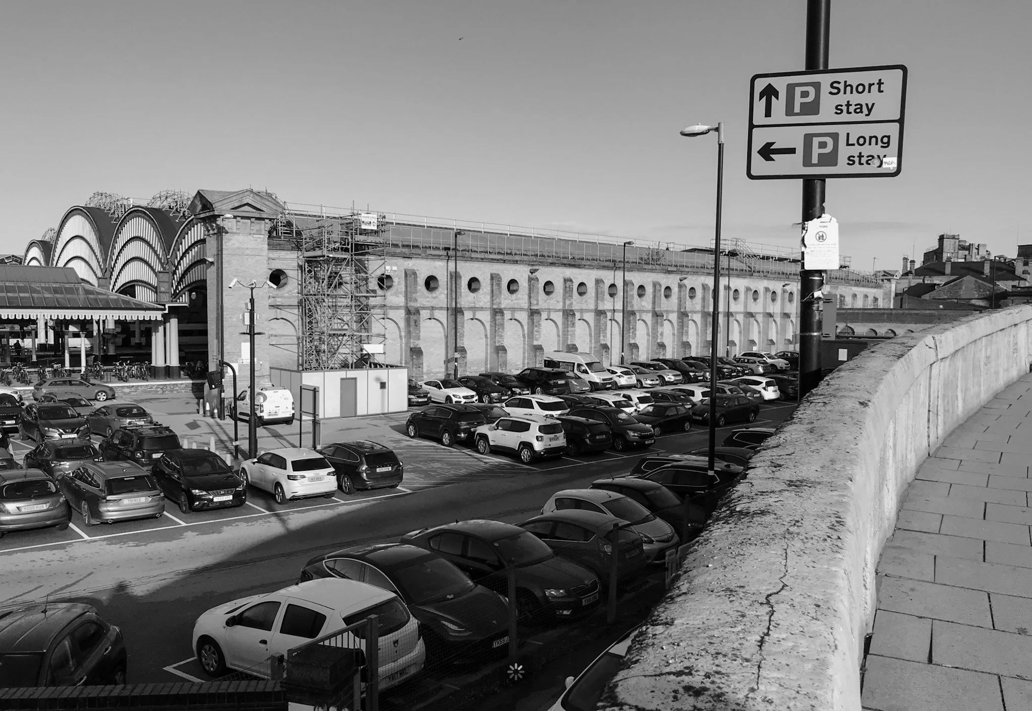 A photograph by Jim Joel of the parking area outside York Station, England, filled with parked cars and framed by the station’s architecture, including a building with distinctive arched windows.