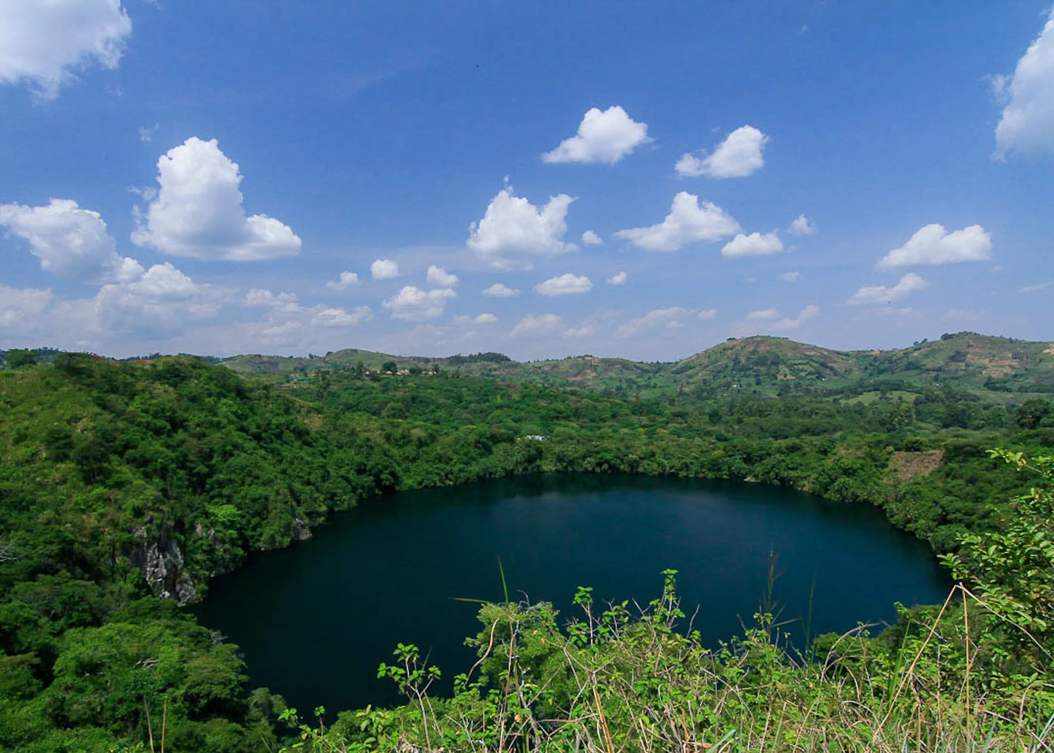 A landscape with a blue sky, scattered white clouds, green rolling hills, and a dark lake or river in the foreground.