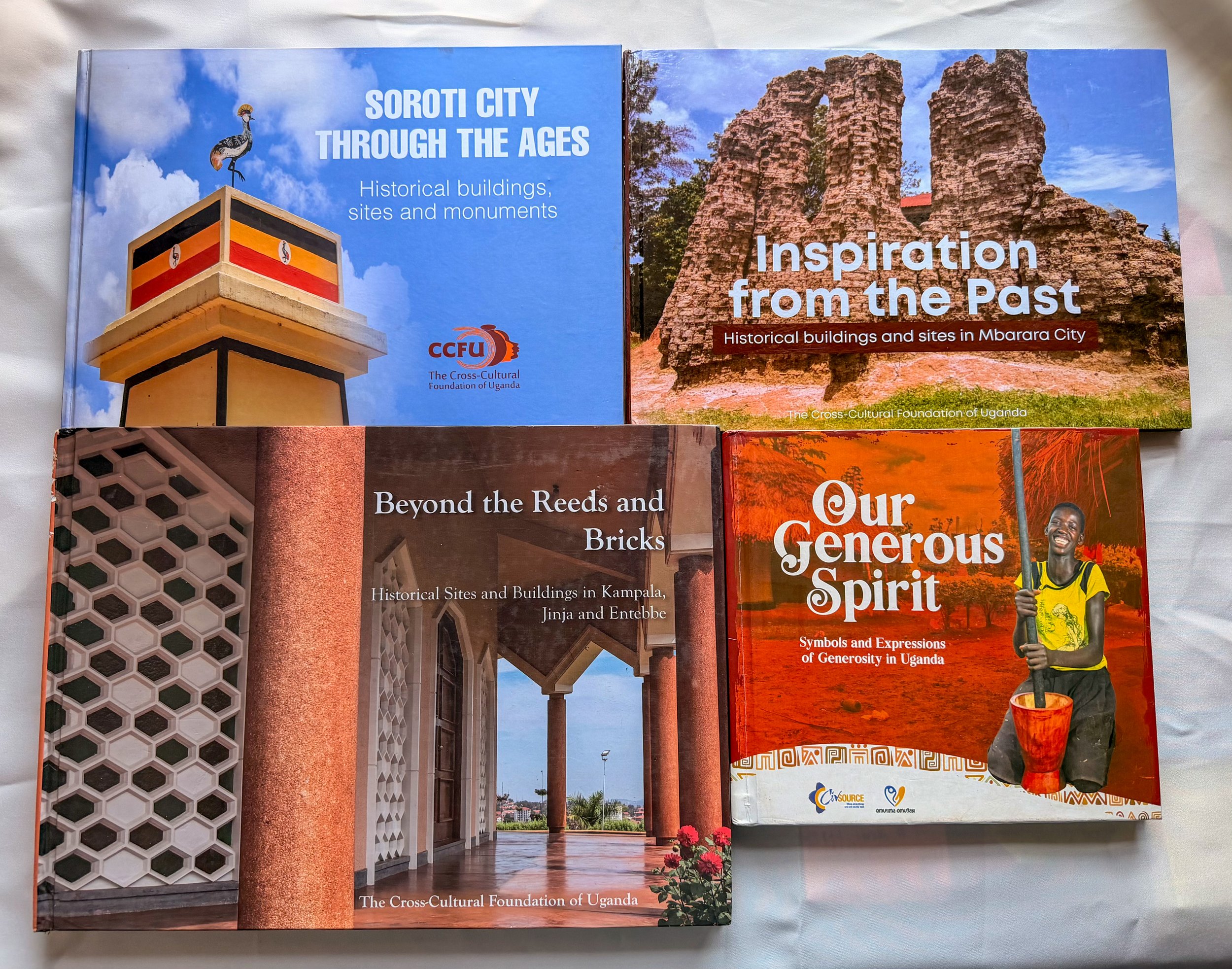 Open book displaying two pages about Uganda's history. The left page features a monument with a bird statue on top, a blue sky with clouds, and text about Soroti City. The right page shows ancient ruins with large stone structures, a blue sky, and te