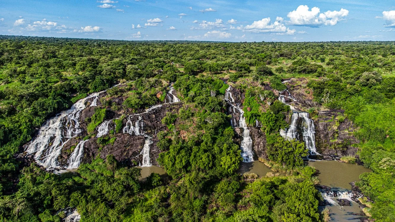 Aerial view of a lush green landscape with multiple waterfalls flowing down rocks into a river, surrounded by dense trees under a partly cloudy sky.