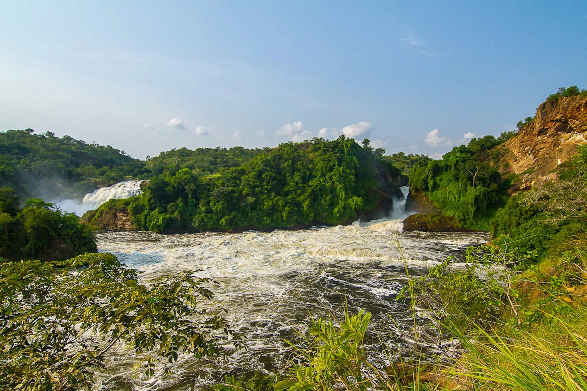 A scenic landscape of a river with a waterfall, lush green trees, and rocky hills under a blue sky with some clouds.
