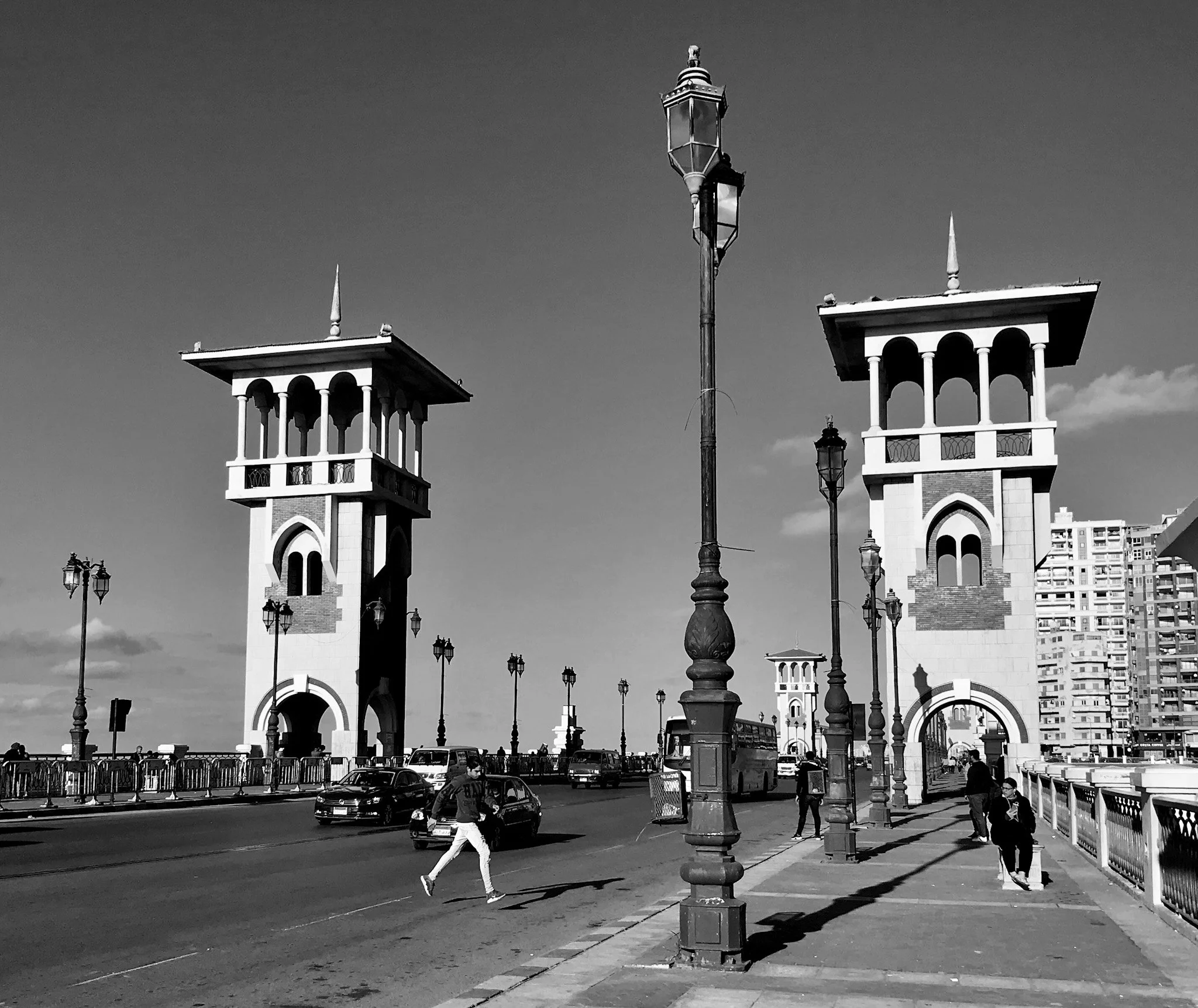 Black and white photo of a city street scene with two towers, street lamps, cars, and pedestrians.