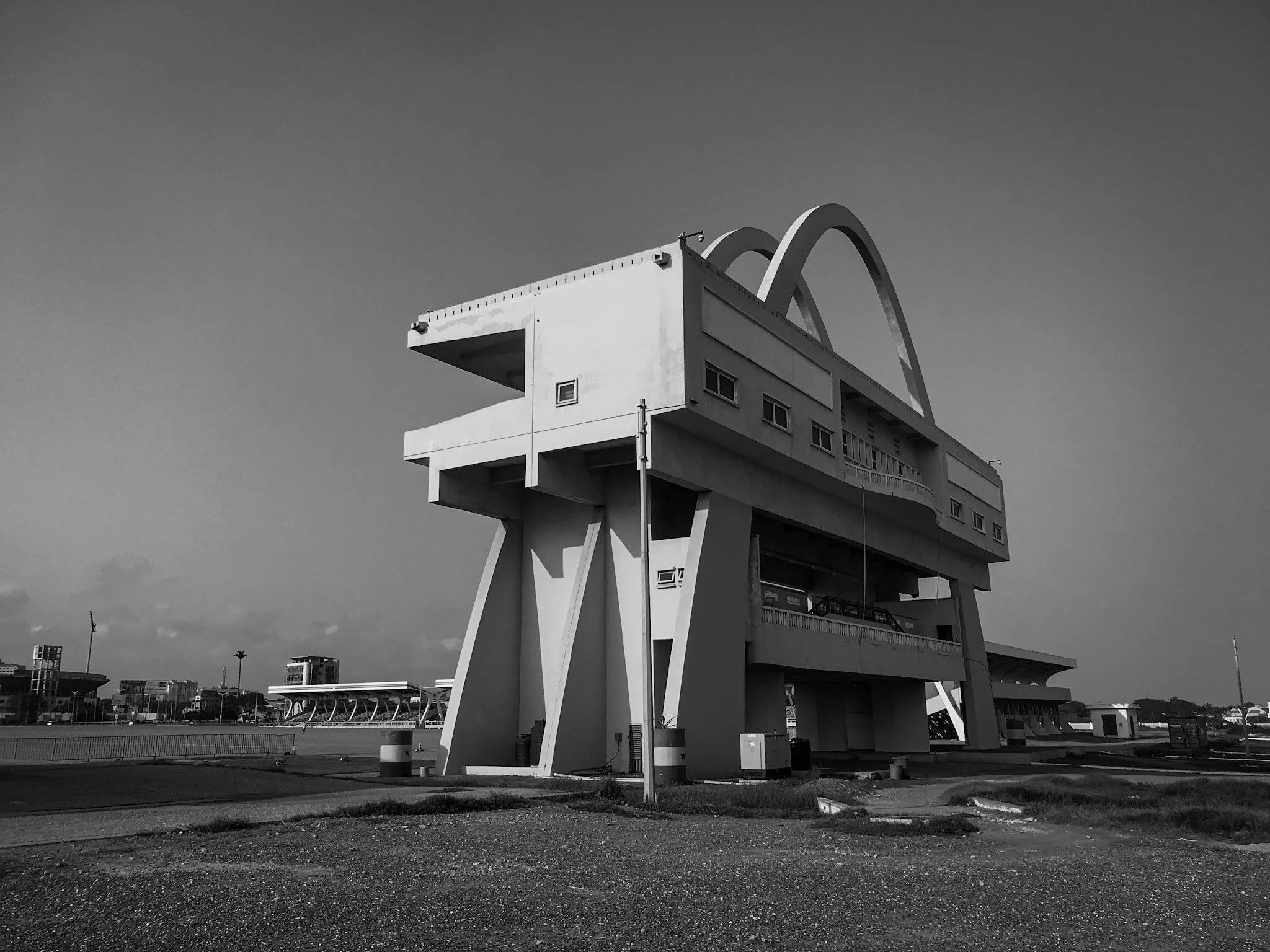 A black-and-white photo of a modern, high-rise building with artistic architectural features, including large curved arches on the upper floors, against a clear sky. Smaller buildings and lampposts are visible in the background and foreground.