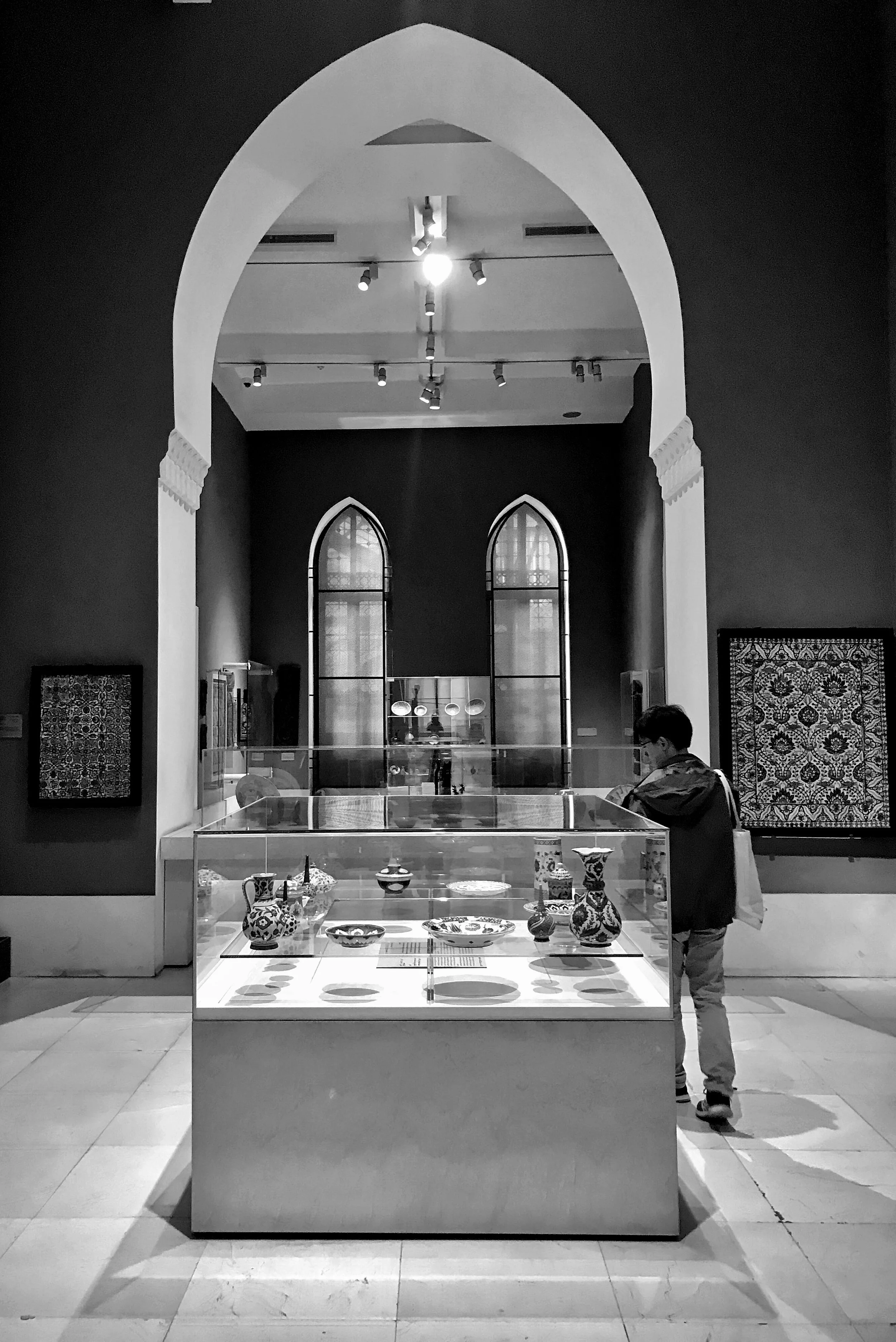 A display case with decorative bowls and vases inside a museum, with a young boy looking at the exhibits. The background features tall, arched windows and artwork on the walls.