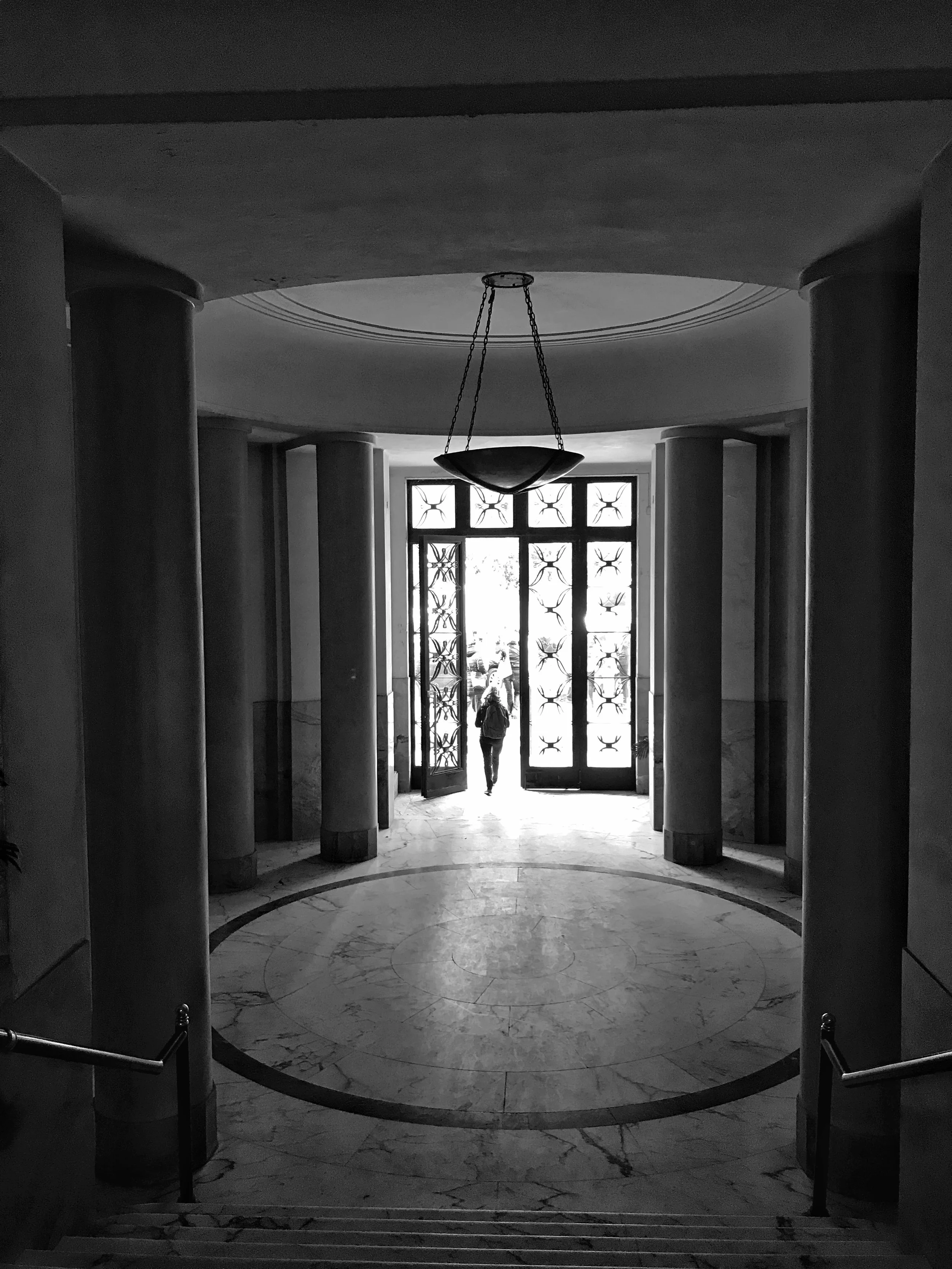 Black and white photo of an interior entrance hallway with marble floors and large columns, showing a person walking through a decorative door with glass panels and patterns. There is a hanging light fixture in the ceiling.