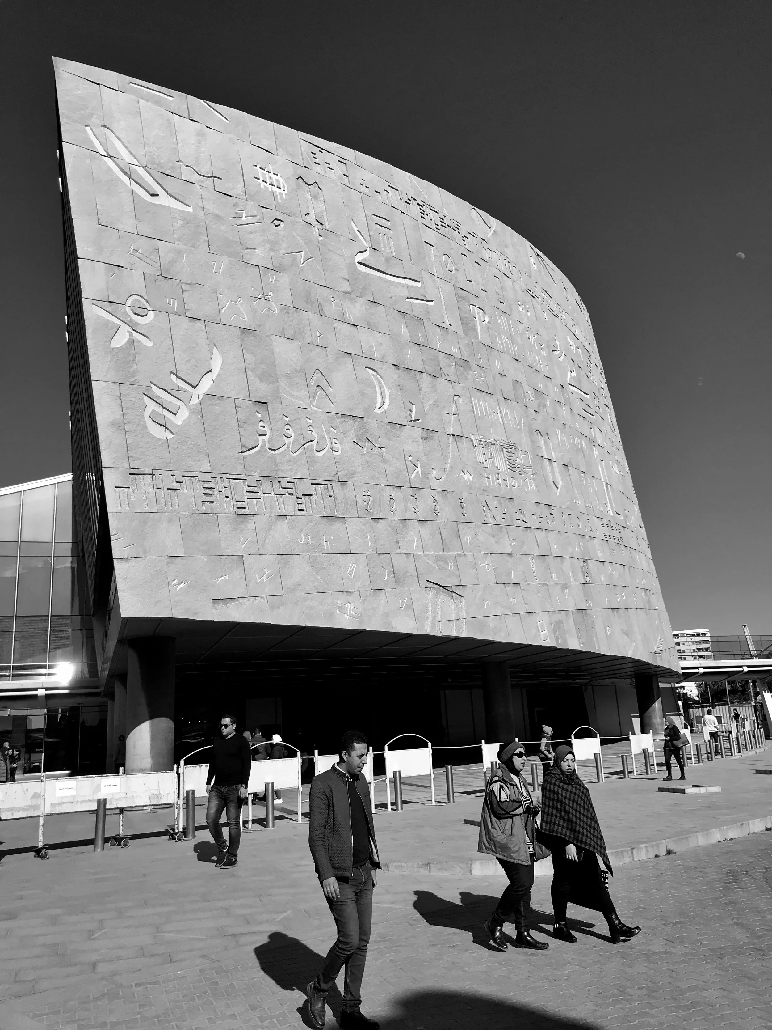A modern building with a curved facade featuring embossed patterns and symbols. People are walking in front of the building, with some waiting behind barriers.
