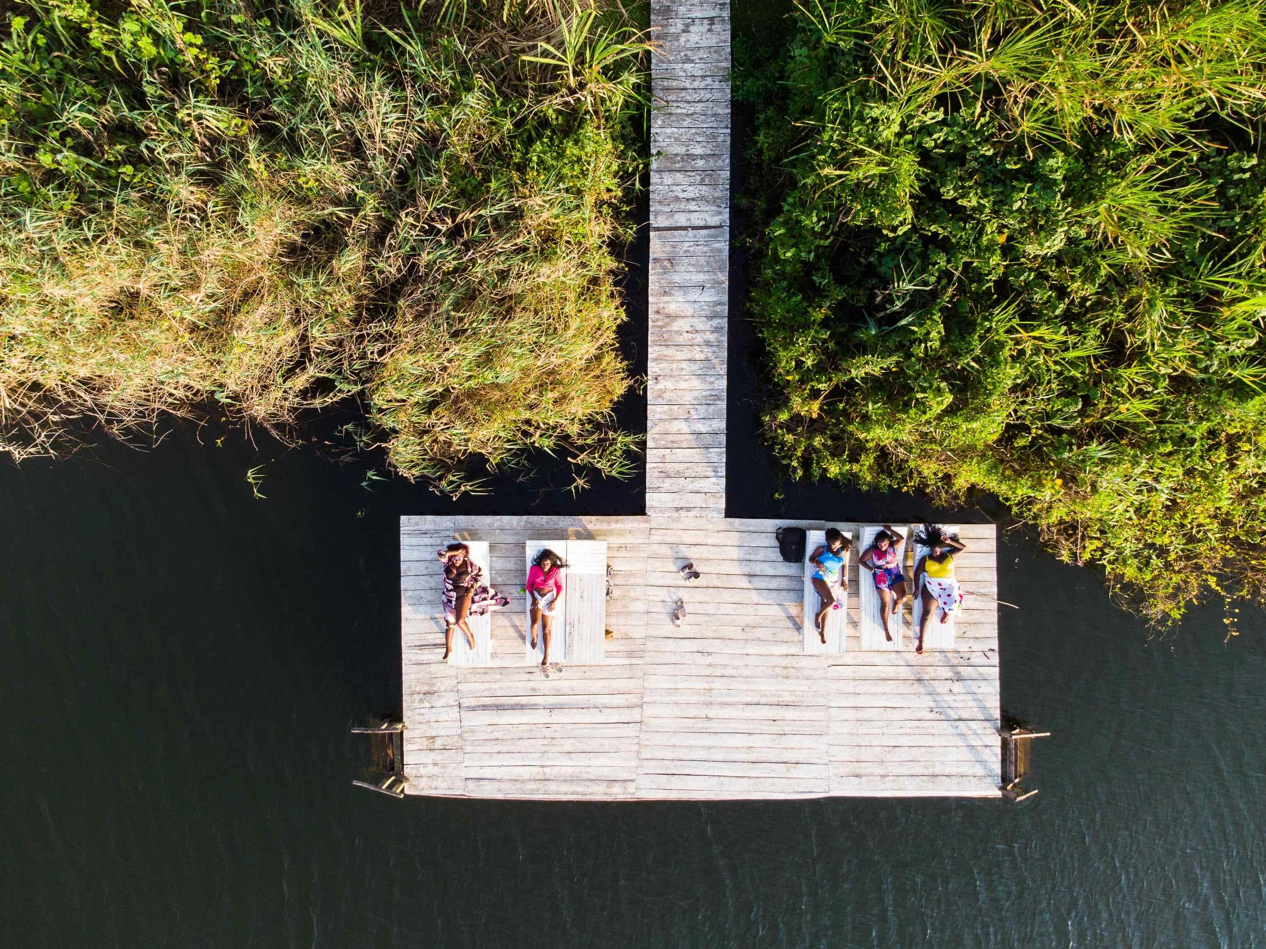 Aerial view of a wooden dock with several people relaxing on lounge chairs, surrounded by green foliage and water.