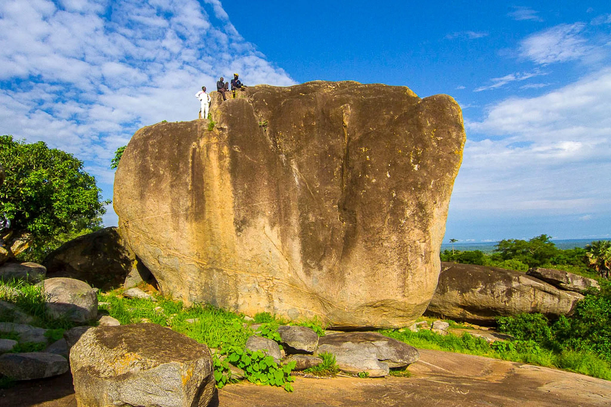 Large rock formation with three people sitting and standing on top, surrounded by smaller rocks and greenery, under a partly cloudy sky.