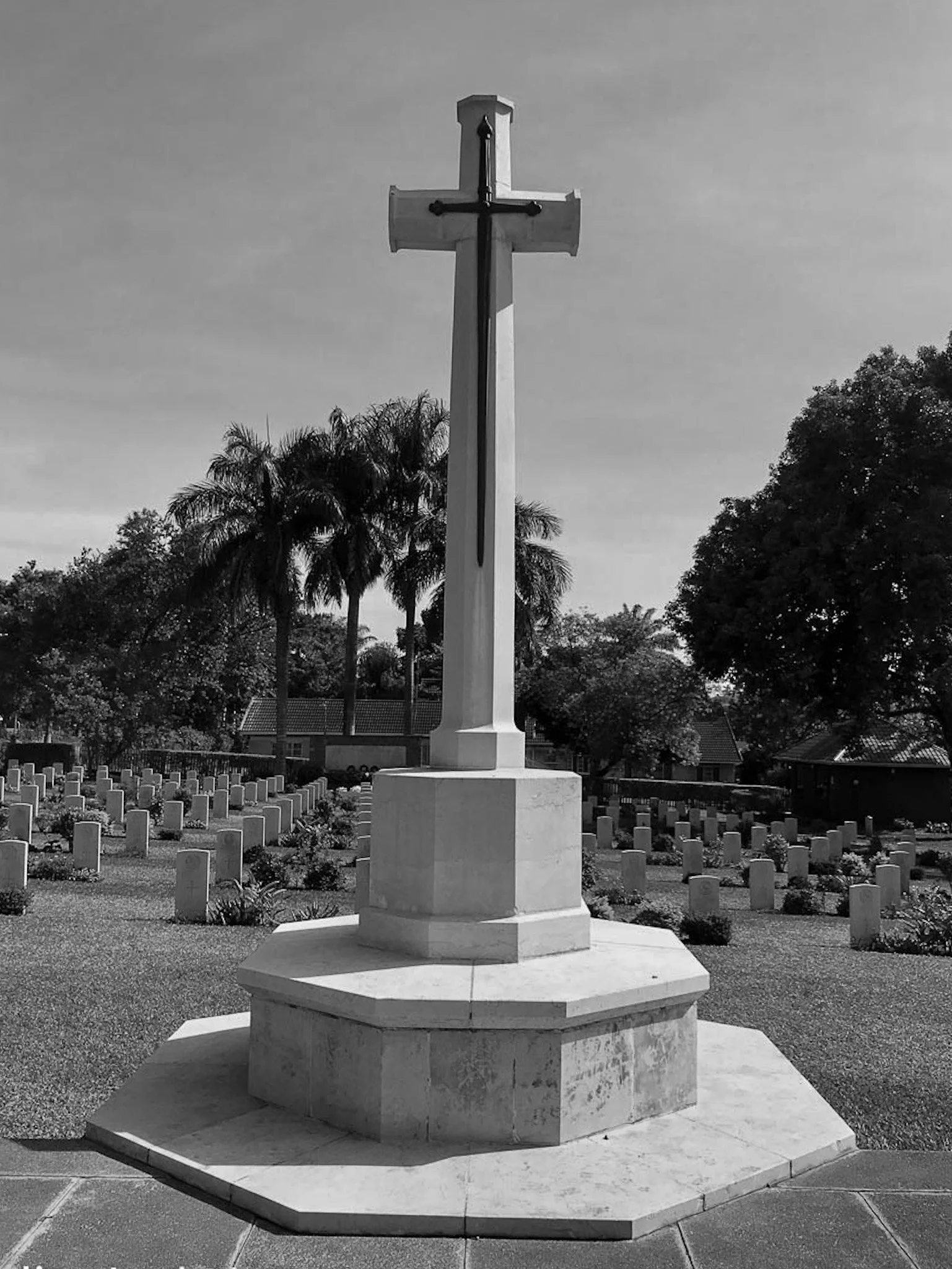 Black and white photo of a large Christian cross monument in a cemetery with rows of grave markers and trees in the background.