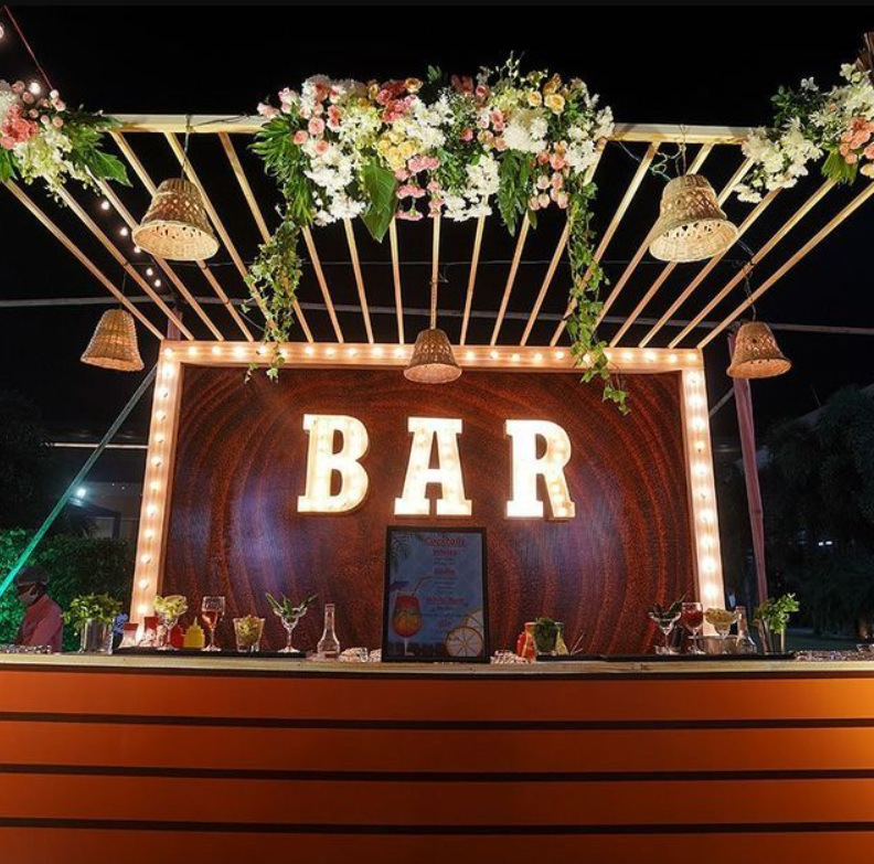 Decorated outdoor bar with illuminated 'BAR' sign, floral arrangements, hanging lights, and wicker pendant lamps. party decoration