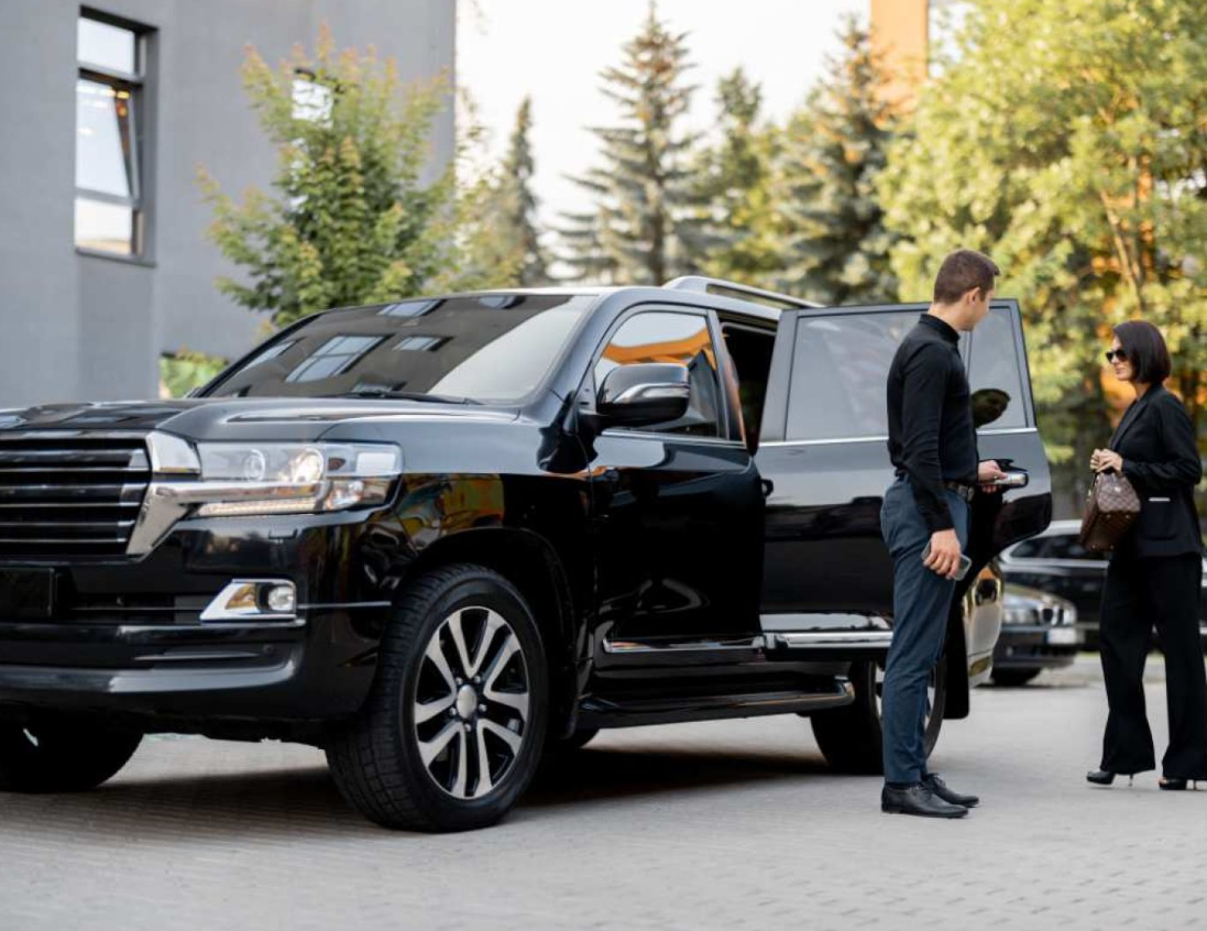 A man and a woman in business attire standing outside near a black SUV with one door open. The background features trees and a building.