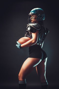 Side view of a female American football player in a helmet, shoulder pads, and gloves, holding a football against a dark background.