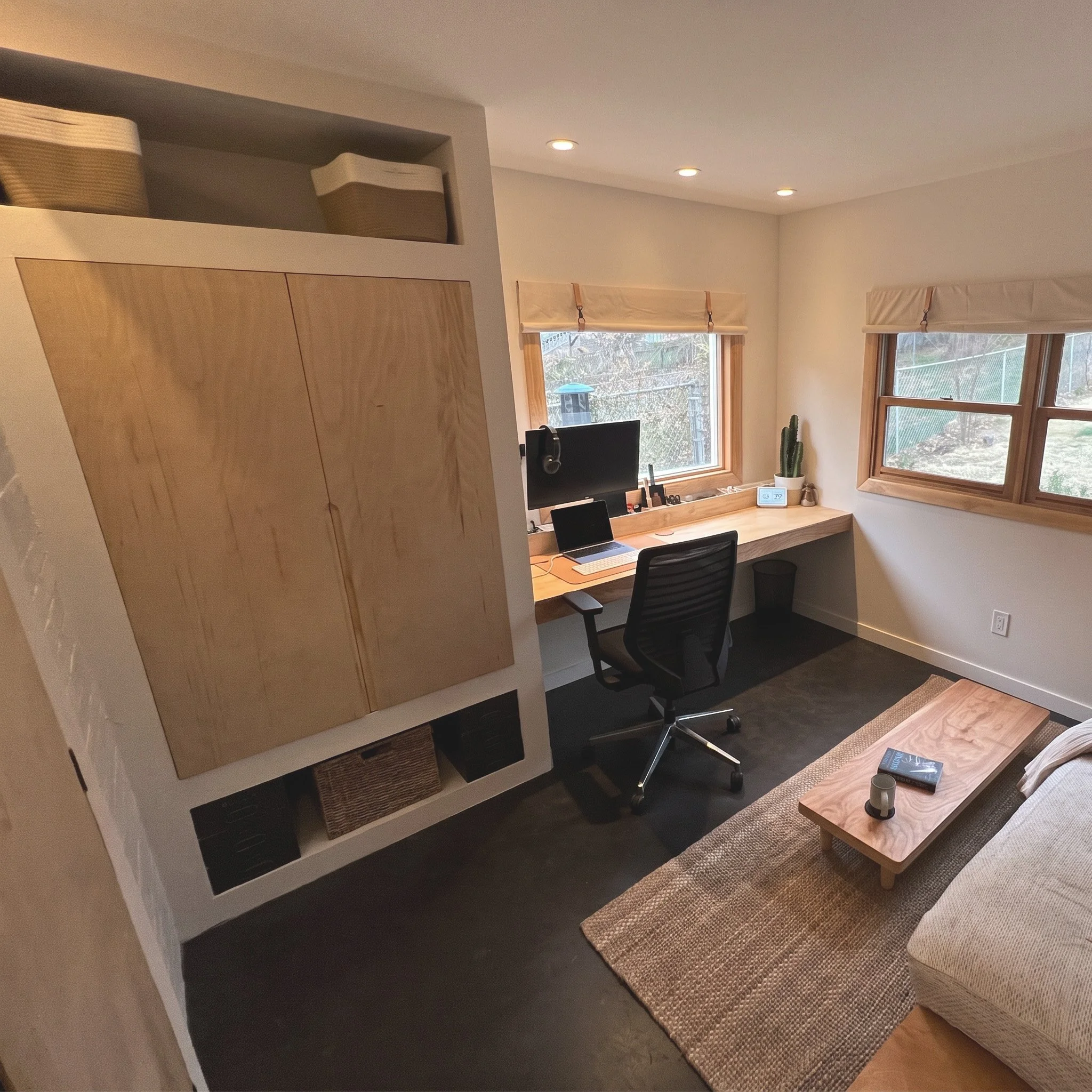 A modern home office with large windows, a wooden desk with a computer monitor, a black office chair, potted cactus, and a beige recliner on a woven rug.