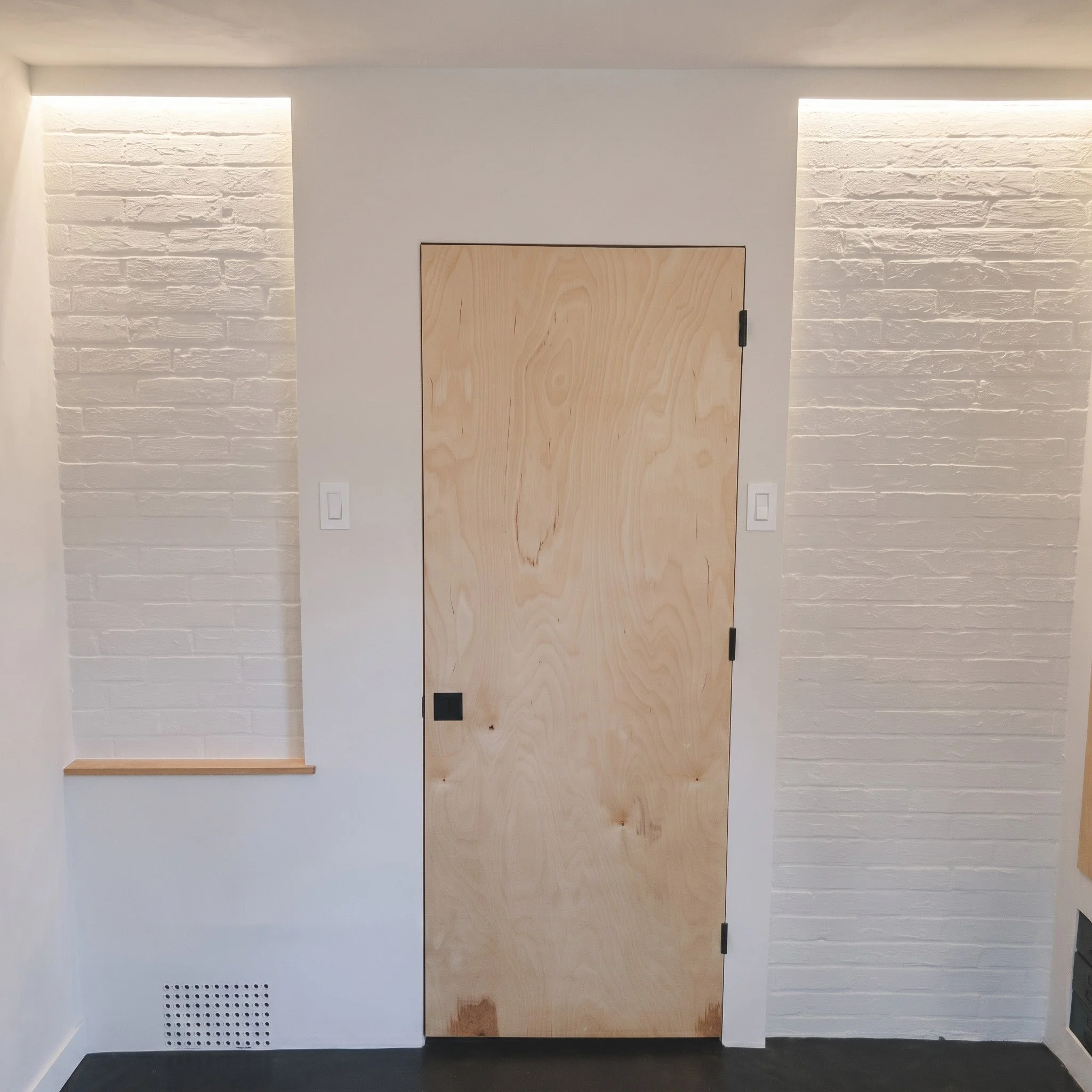 Interior wall with two recessed lighting fixtures, a wooden door, a white wall with a vent, and a black floor.