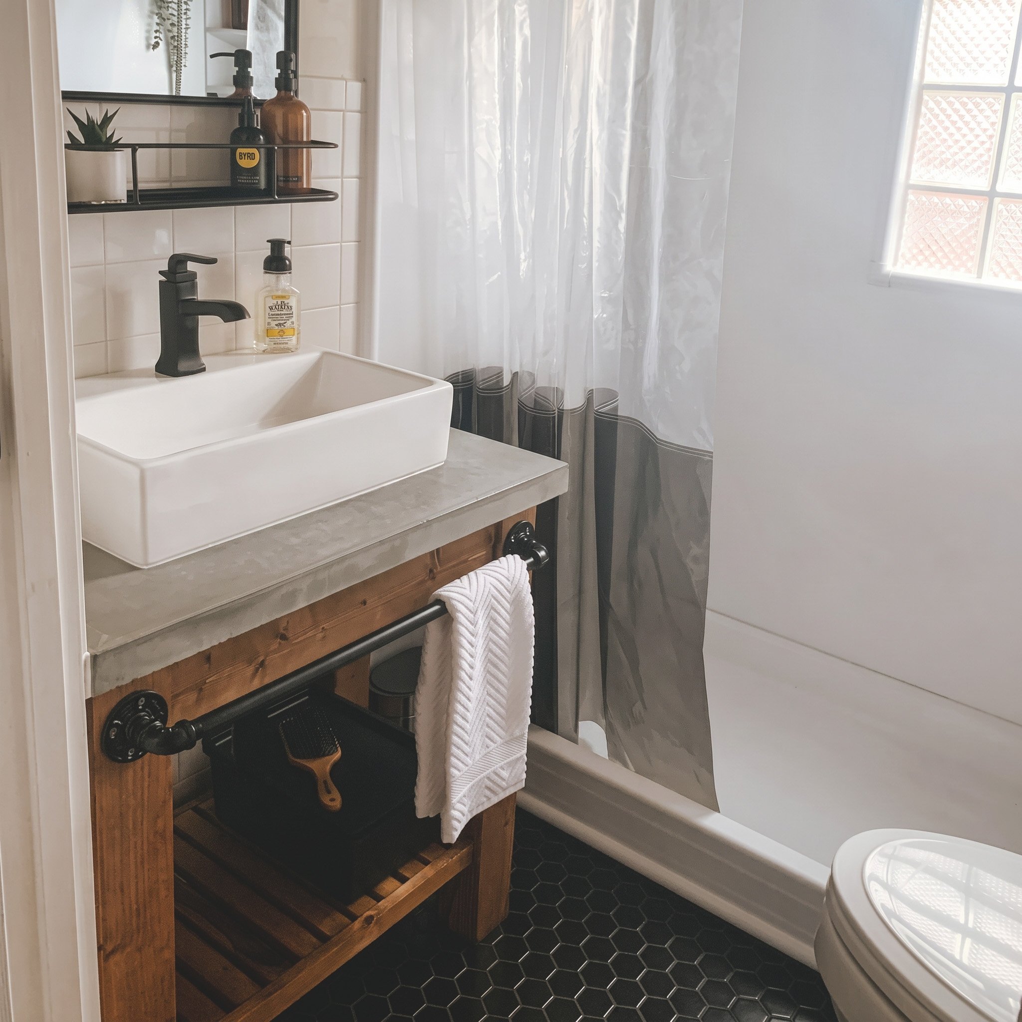 A small bathroom with a white sink, black faucet, and a wooden vanity with a slatted bottom shelf. Above the sink is a black wall-mounted shelf holding bottles and a small potted plant. There is a mirror above the shelf. To the right, a shower curtain with a subtle pattern hangs in front of a bathtub. The floor has black hexagonal tiles, and sunlight is coming through a window with frosted glass blocks.