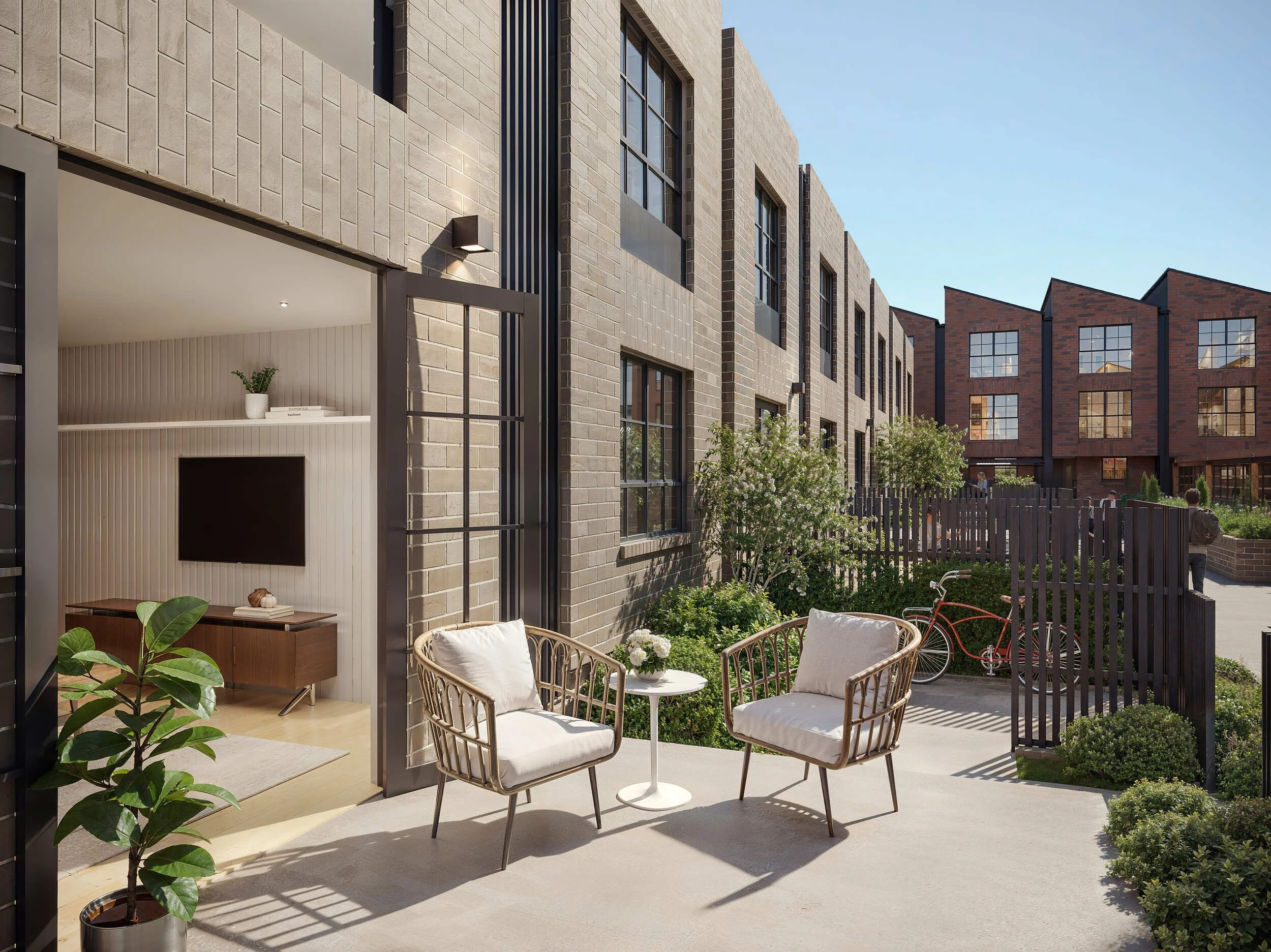 Modern apartment building with a small outdoor patio area, featuring white and beige furniture, green plants, and a black metal fence, with other buildings and trees visible in the background on a clear day.
