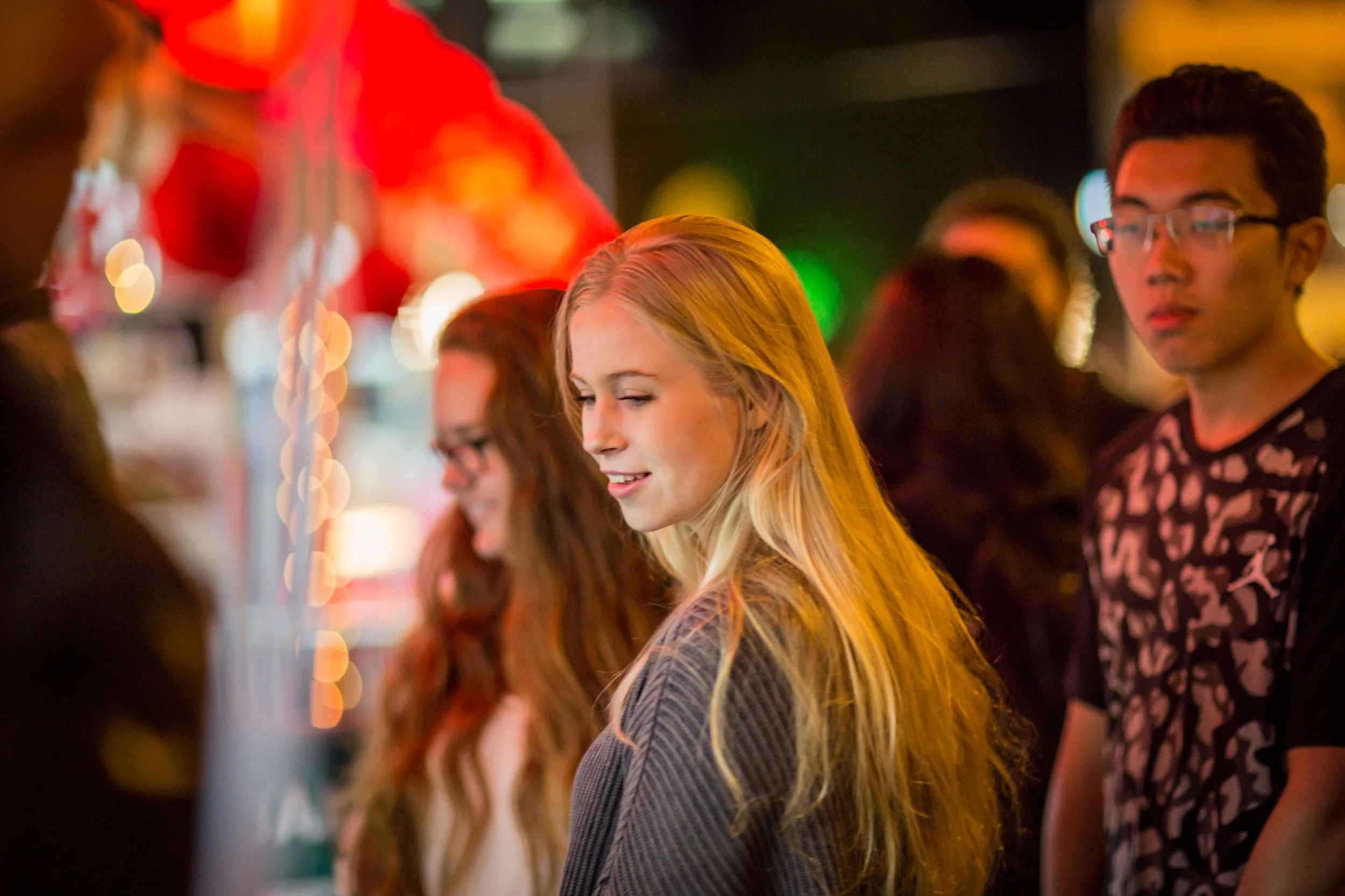 People standing in line at a colorful nighttime event decorated with red and yellow balloons, with blurred background lights.