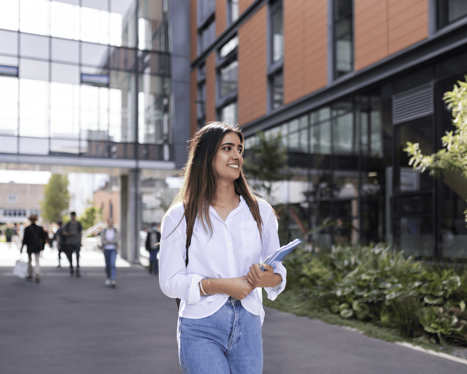 Young woman with long brown hair, wearing a white shirt and blue jeans, holding notebooks, standing outside modern office building with glass and wood exterior, other people walking in the background.