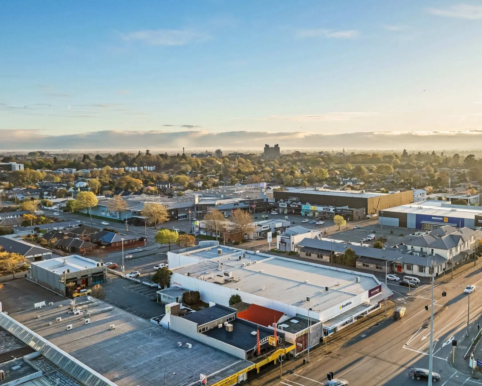 Wide aerial view of a commercial area with retail stores, parking lots, and roads. Residential houses and a distant city skyline are visible in the background, with a sky full of light clouds.