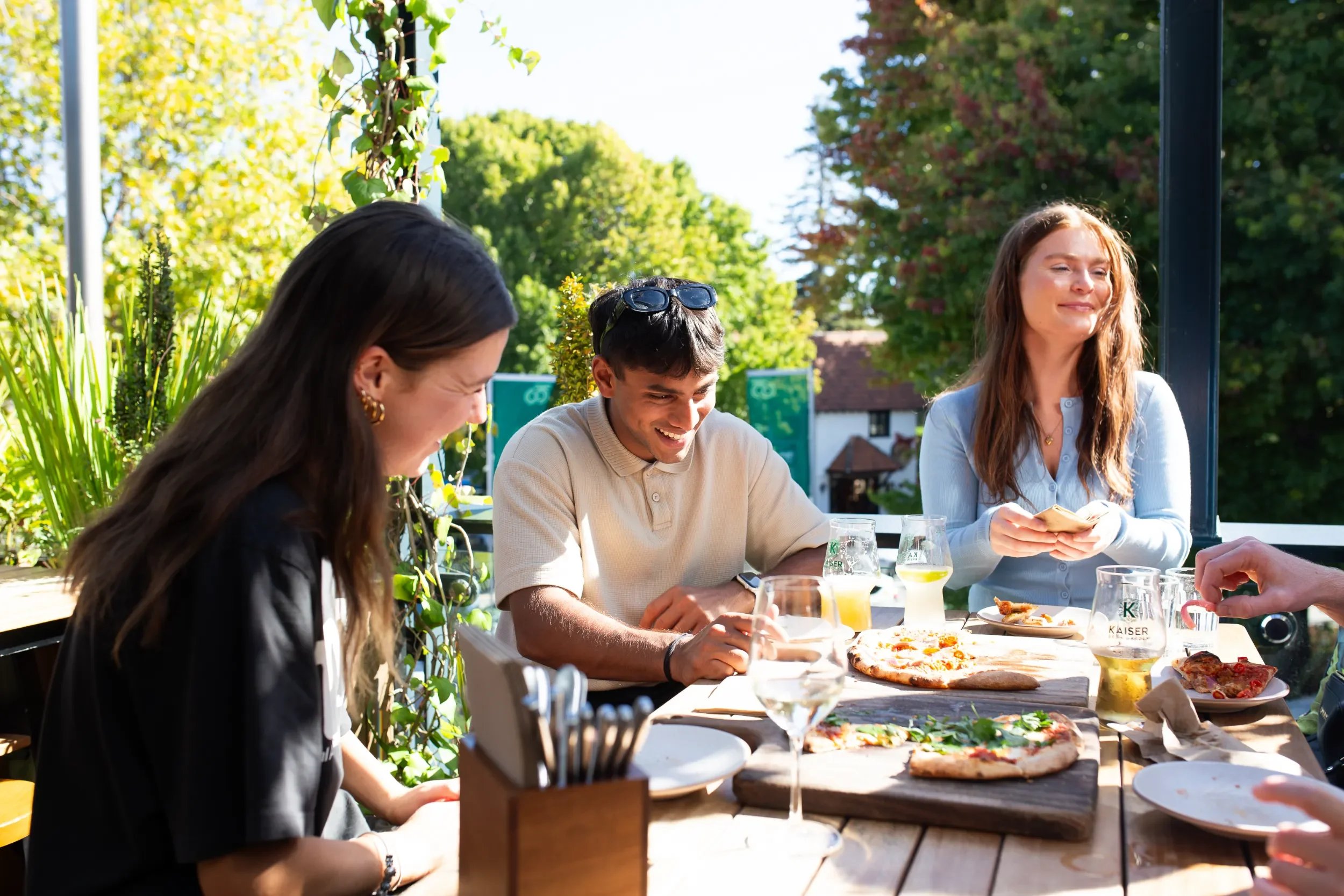 A group of people gathered around a table outdoors enjoying a meal on a sunny day. There are glasses of drinks and pizza on the table.