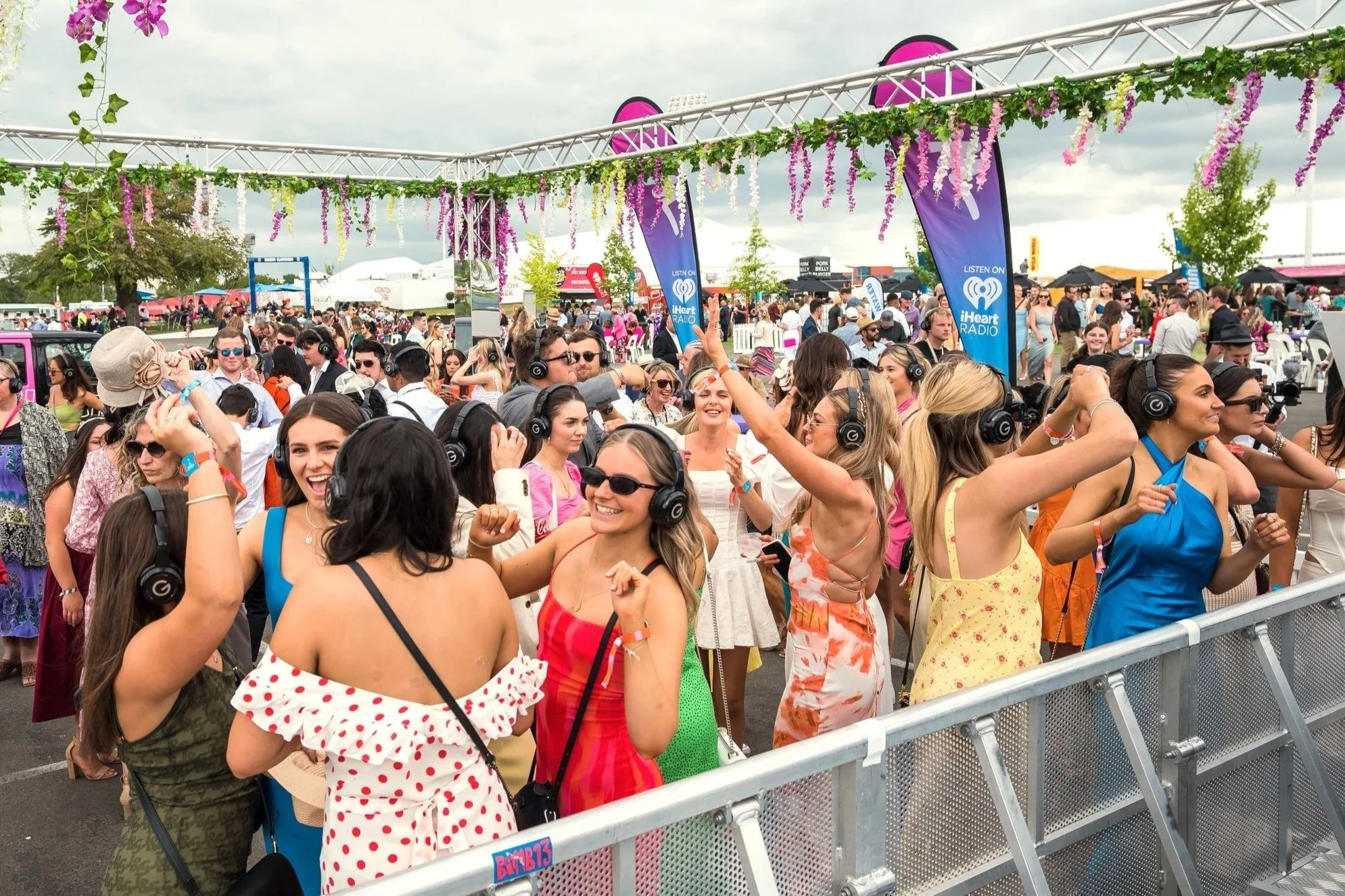 People attending an outdoor music festival, many wearing headphones and colorful summer clothing, with a decorated border overhead, and flags promoting iHeartRadio.