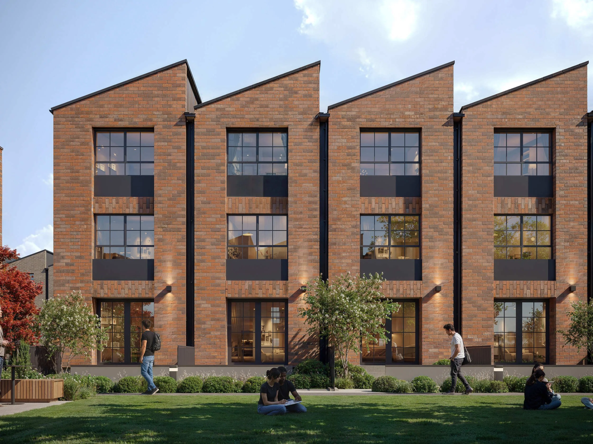 A modern brick apartment building with large black-framed windows and a sloped roof, set against a blue sky with a few clouds. People are sitting and walking in a landscaped courtyard with grass, trees, and shrubs.