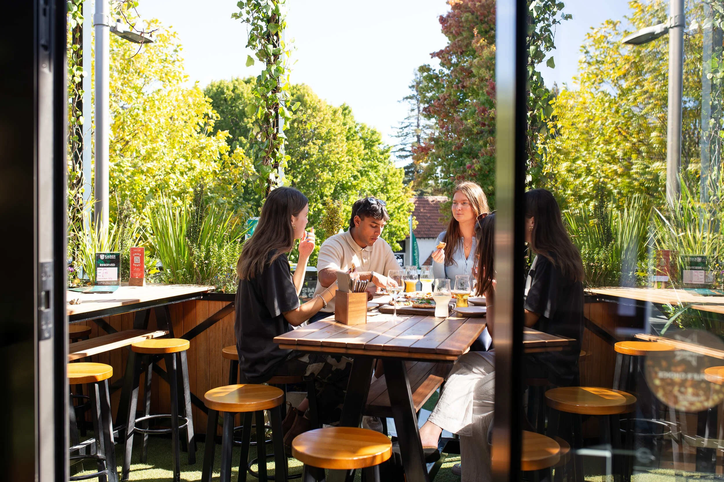 A group of five young adults sitting around a wooden table, eating and talking outside at a restaurant on a sunny day, surrounded by green trees and plants.