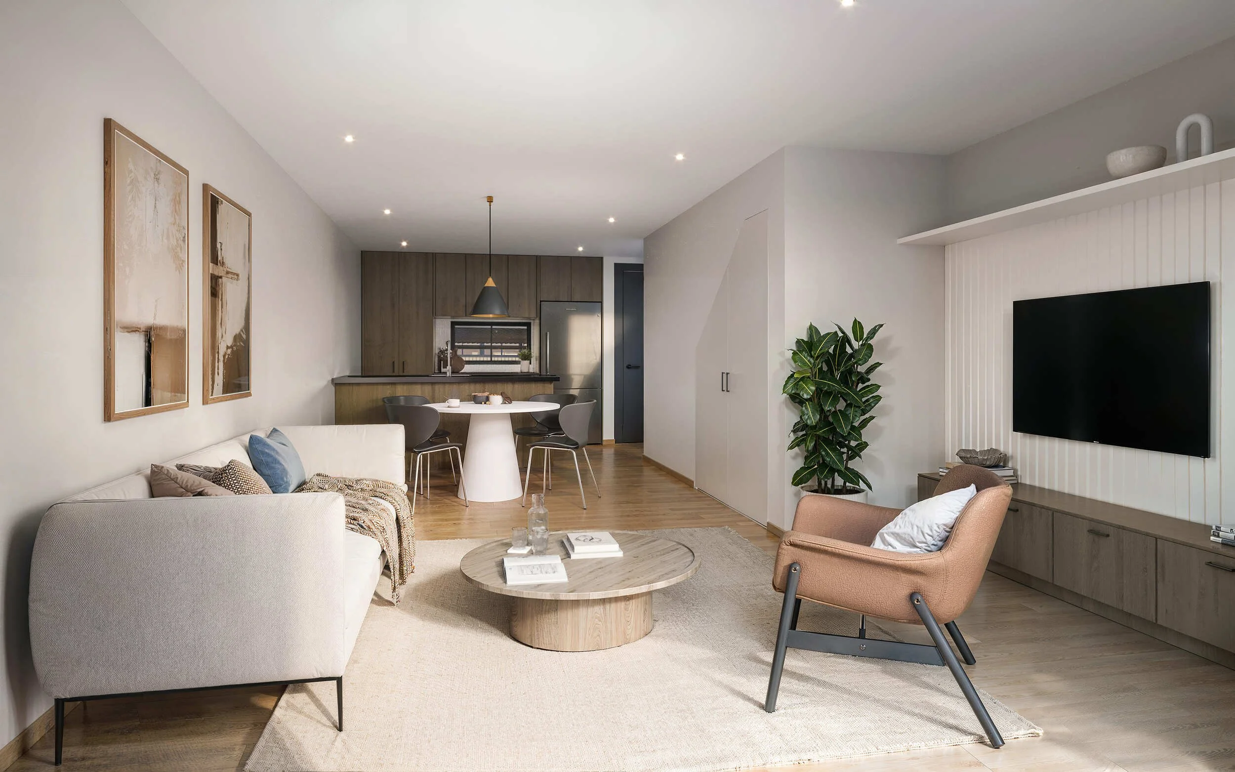 Modern living room with white sofa decorated with pillows, a round wooden coffee table, and armchair, with a large green plant, wall-mounted TV, and artwork on the wall. Open kitchen with wooden cabinets and dining area in the background.