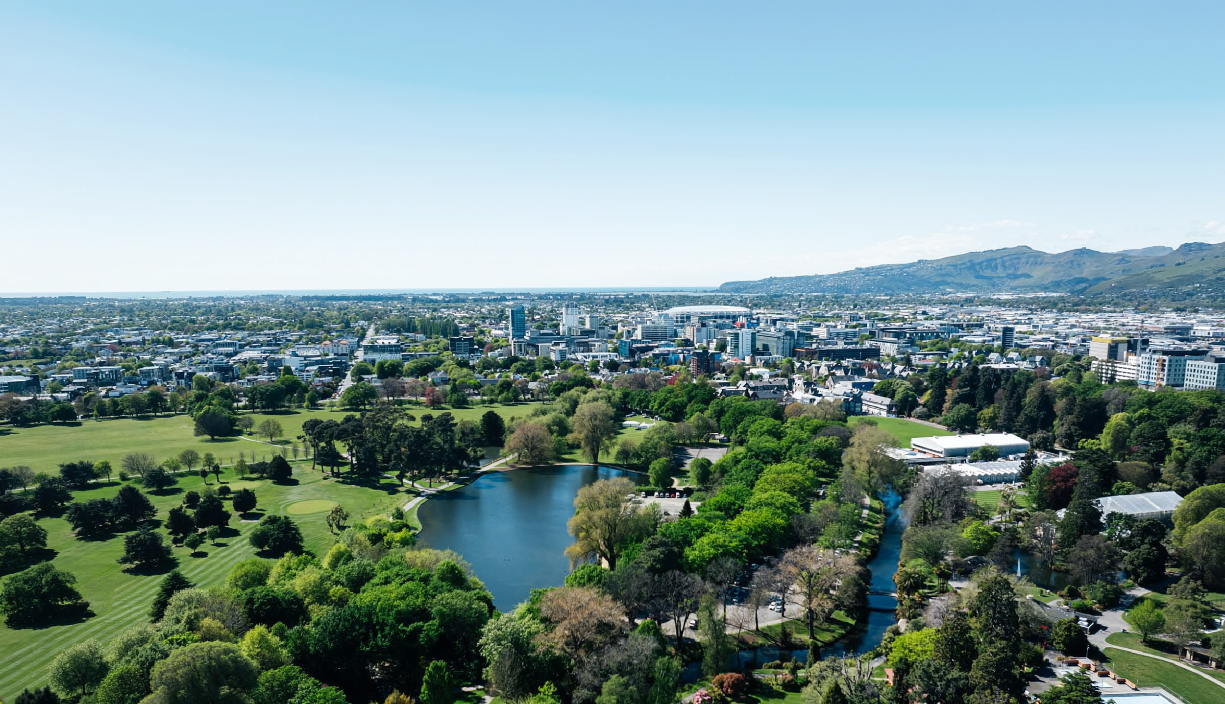 Aerial view of a city skyline with high-rise buildings, a large park with a pond, trees, and a mountain in the background under a clear blue sky.
