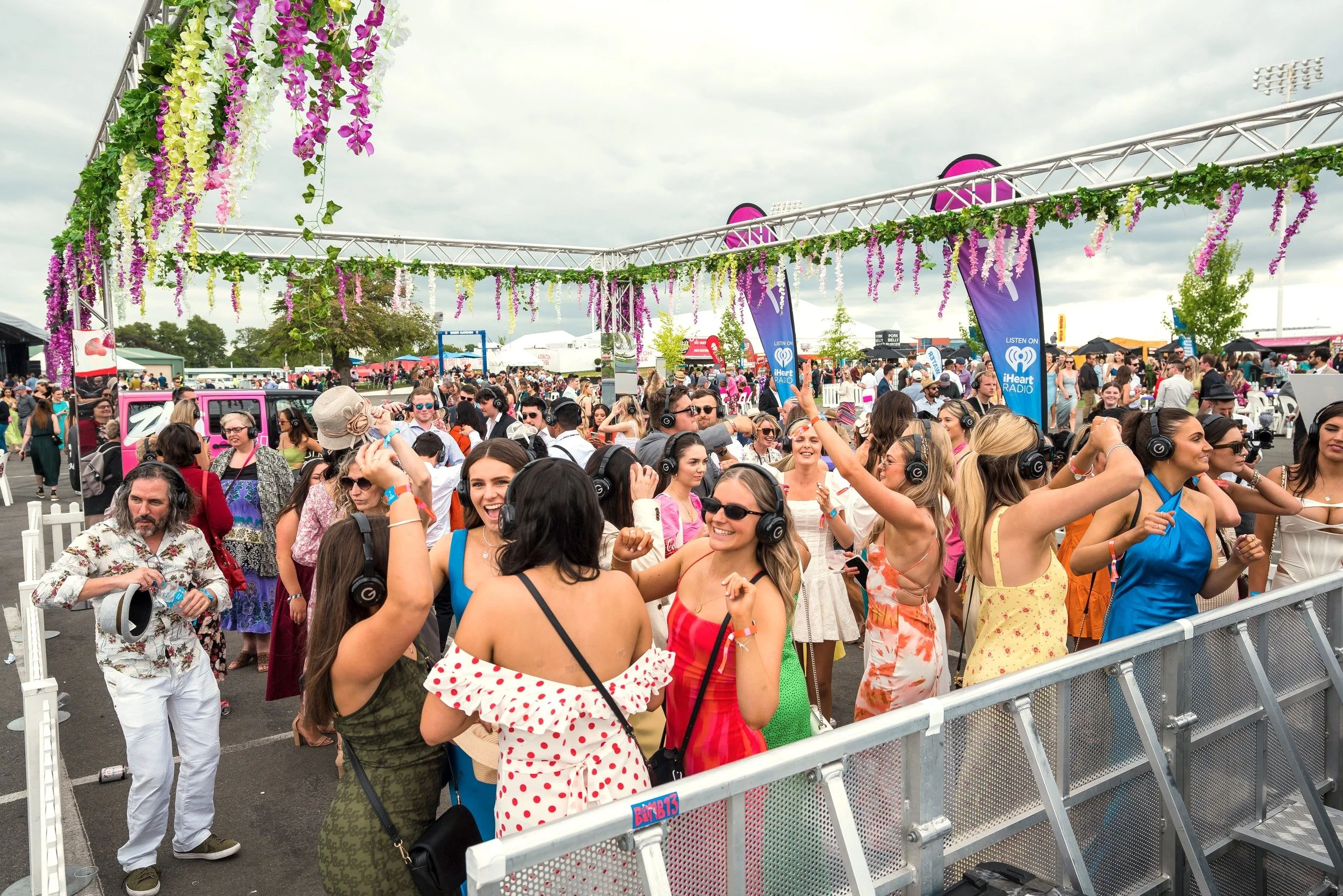 Crowd of people at an outdoor festival, many wearing headphones, with tents, banners, and floral decorations, under cloudy skies.