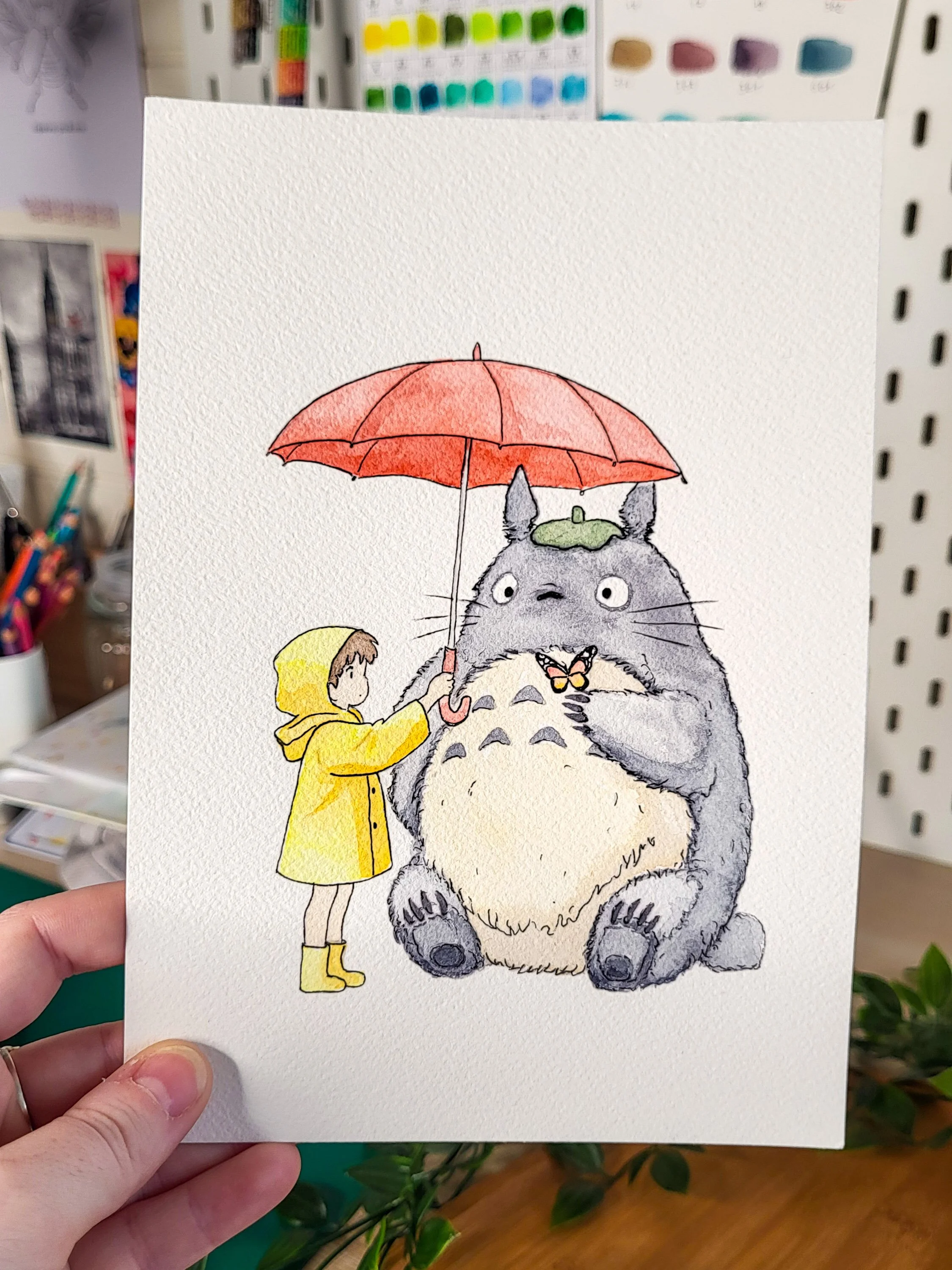 Une illustration d'une fille en manteau jaune tenant un parapluie au-dessus d'un personnage, Totoro, assis sur le sol. Totoro est un grand creature gris avec un ventre blanc, deux oreilles pointues et une expression curieuse, et porte un chapeau vert